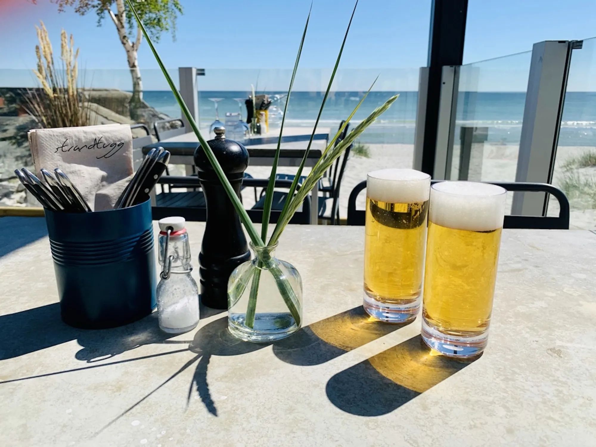 Två Falsterbo Longdrink-ölglas på ett restaurangbord vid havet med pepparkvarn, saltkar och utsikt över stranden.