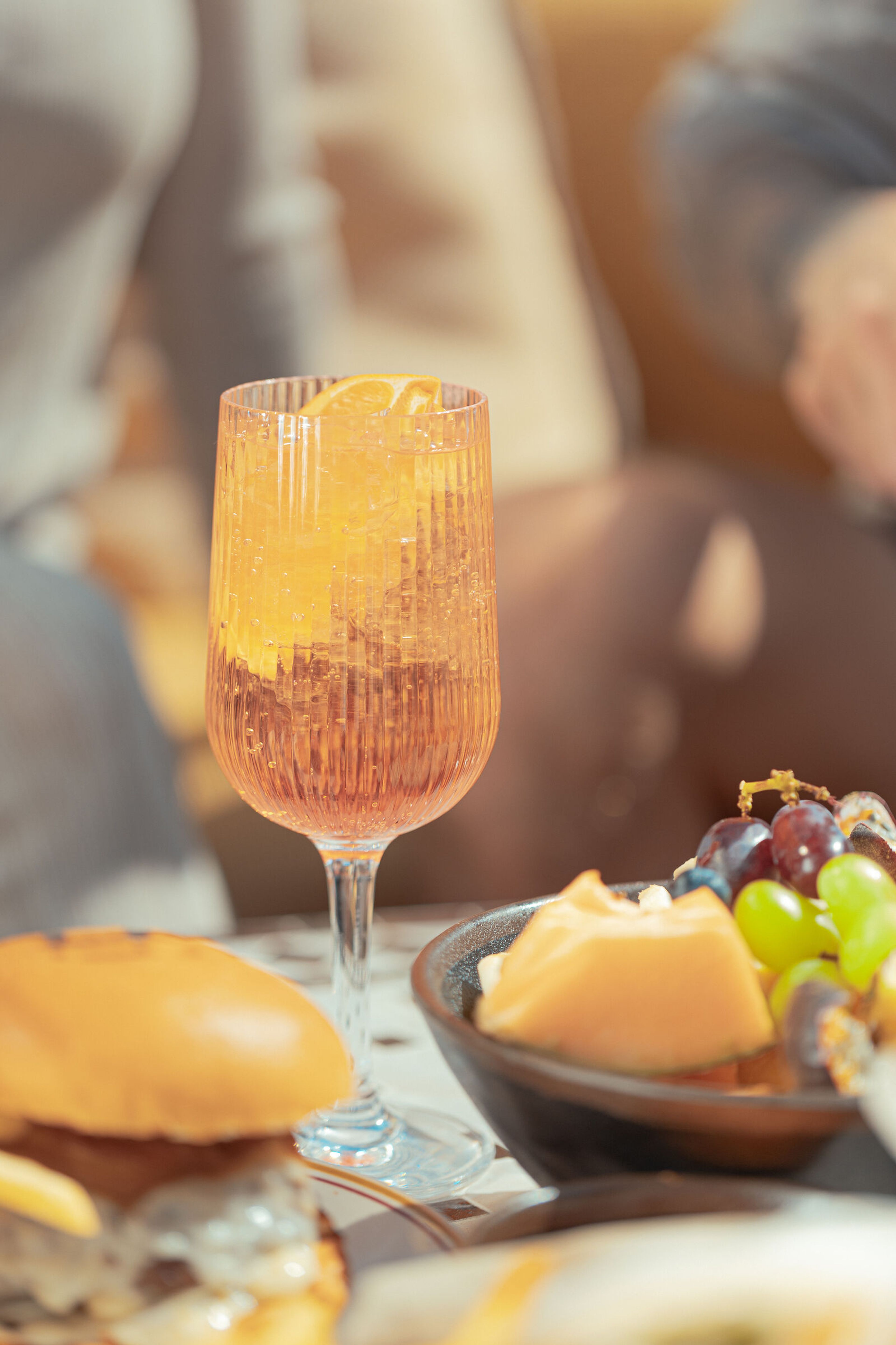 Räfflat vinglas med en orange cocktail och citrusskiva på ett utomhusbord bredvid en skål med frukt och ost.