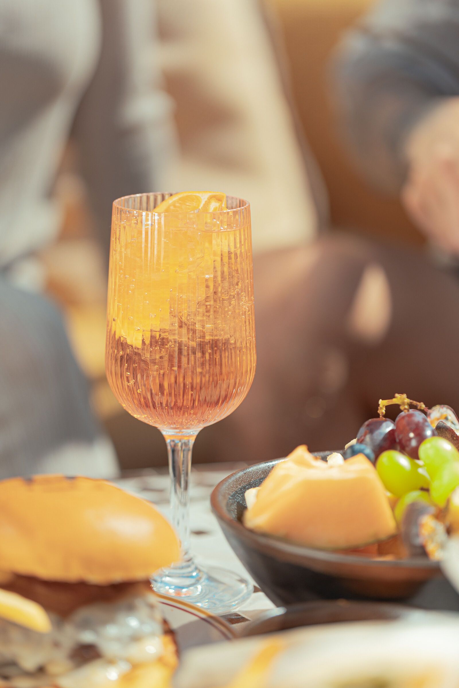 Räfflat vinglas med en orange cocktail och citrusskiva på ett utomhusbord bredvid en skål med frukt och ost.