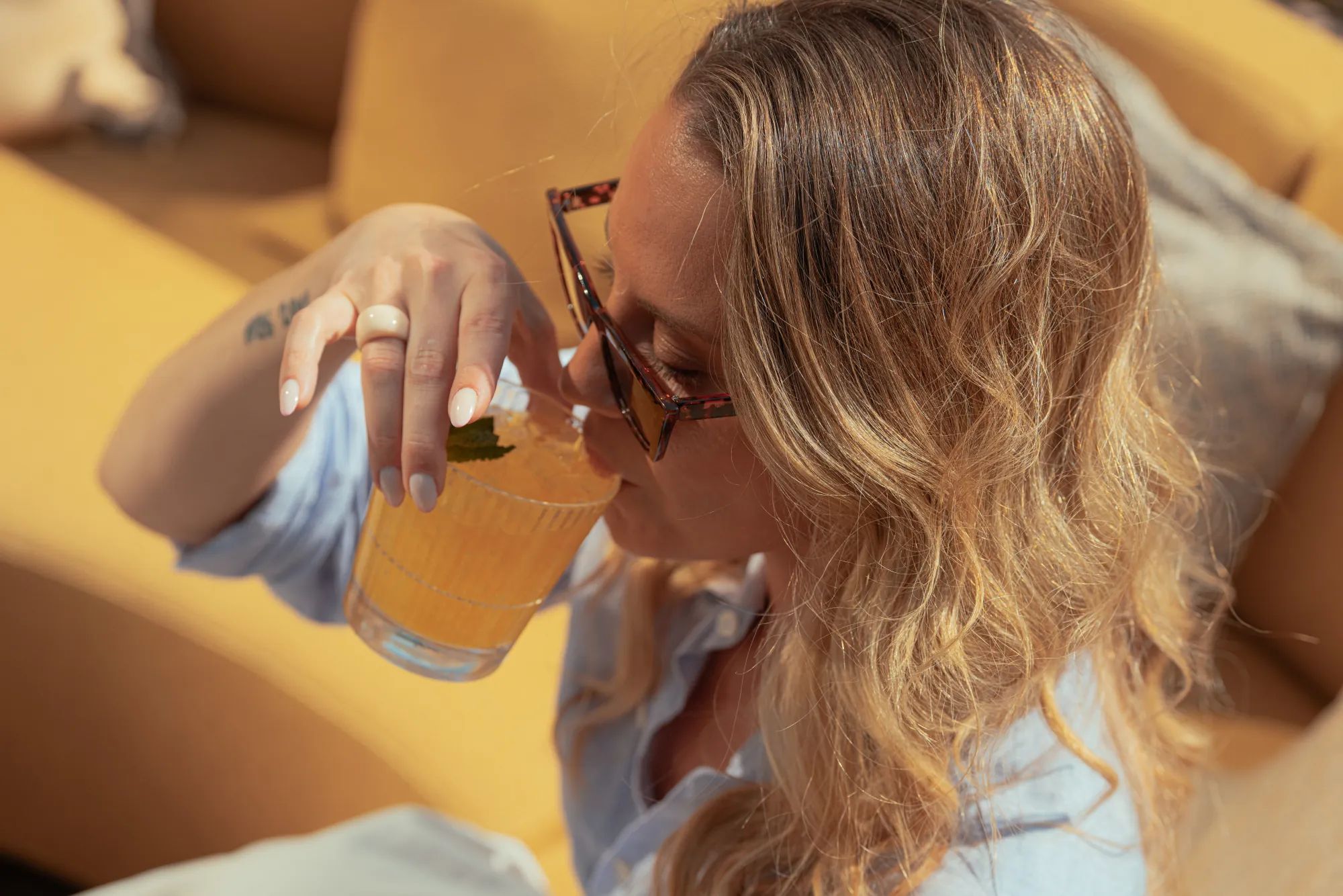Kvinna med solglasögon smuttar på en cocktail ur ett Stripe Rocks-glas i en solbelyst gul soffa.