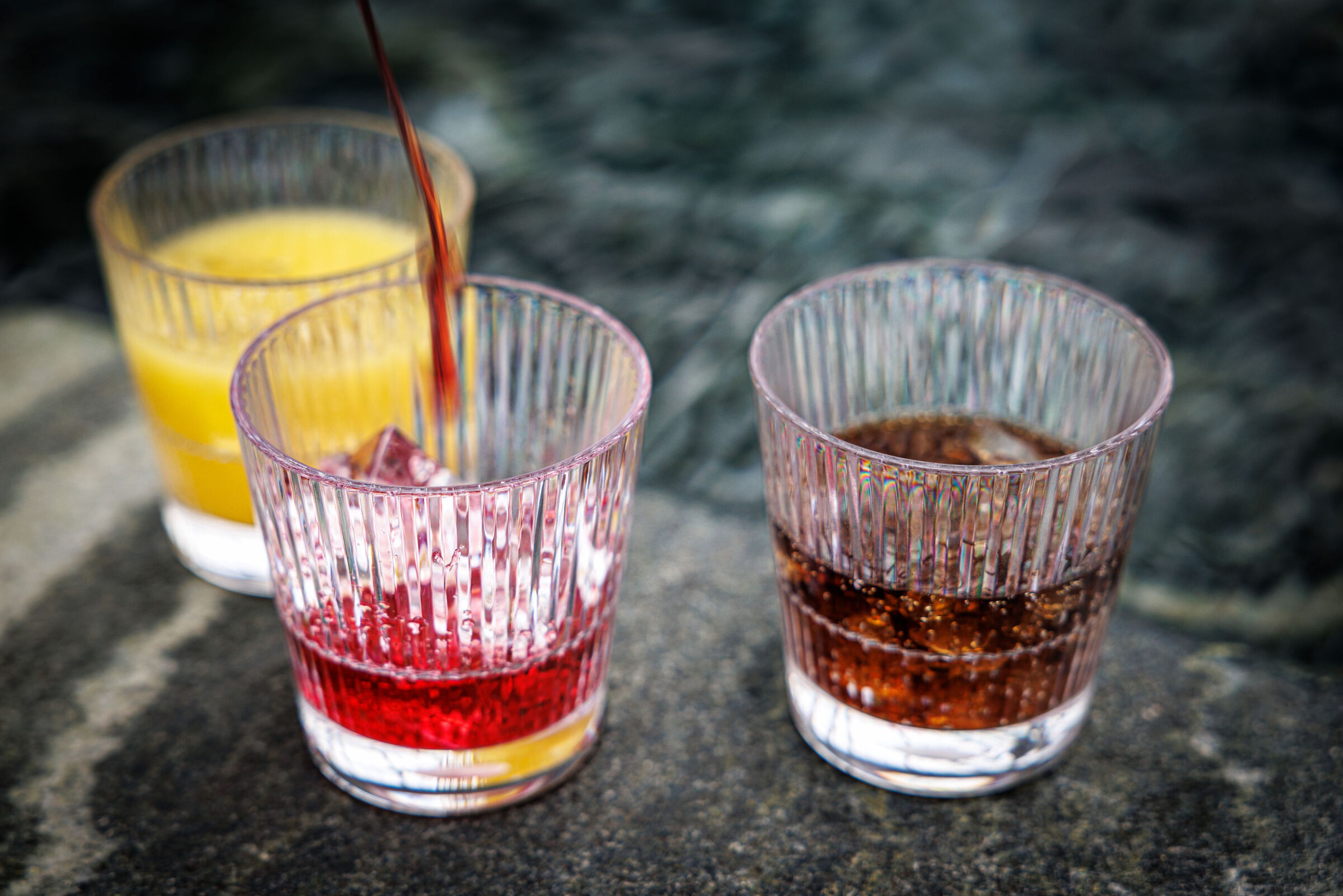 Räfflade tumblerglas med olika drycker, inklusive röd, gul och mörk läsk.