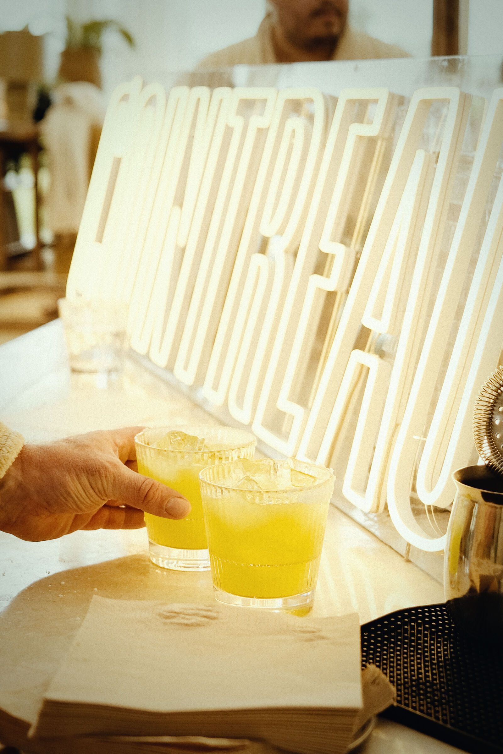 Hand som håller en burgare bredvid en orange cocktail i ett räfflat glas på ett bord.