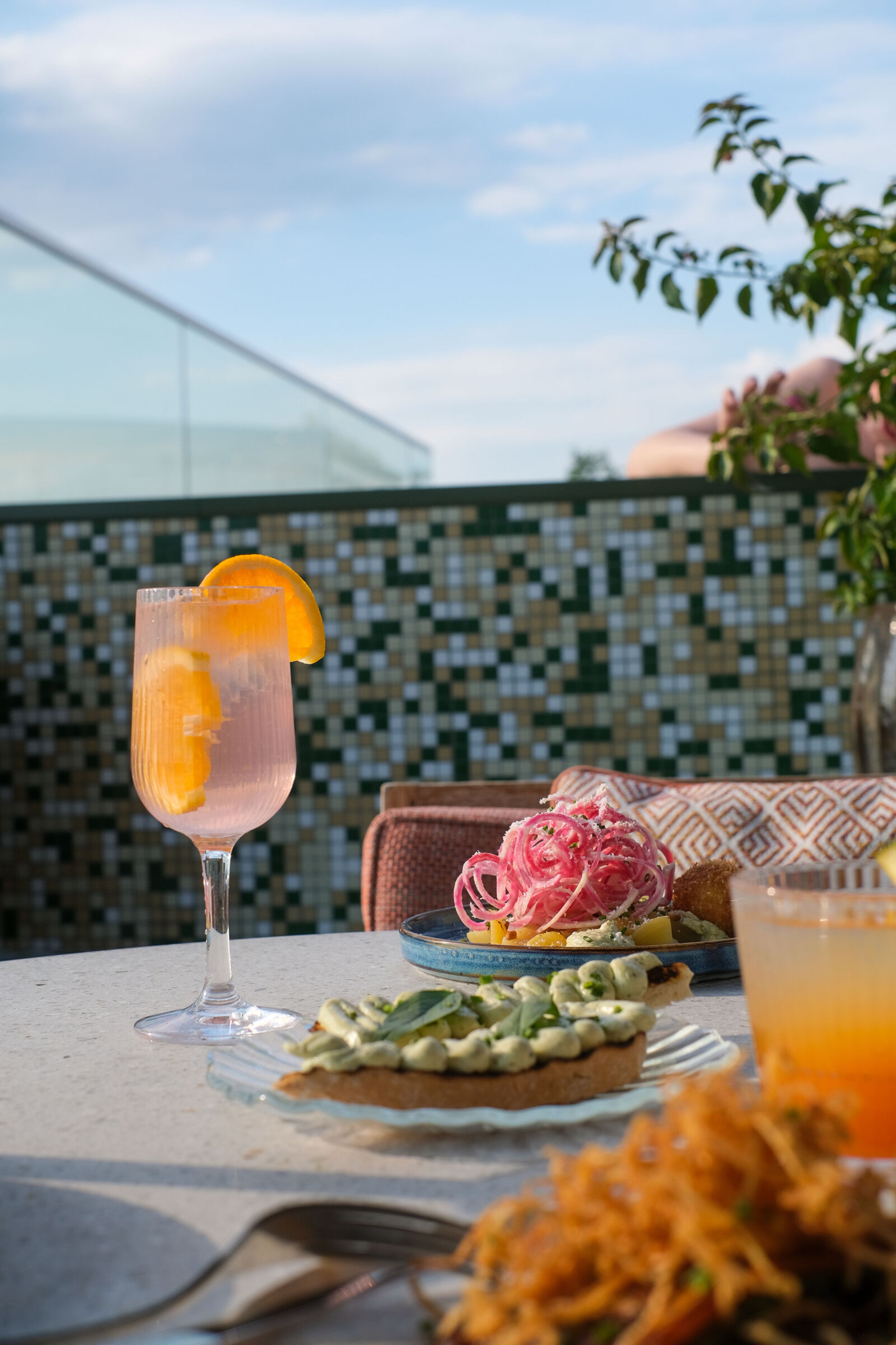 Uteservering med förrätter och cocktails serverade i räfflade glas, inklusive ett vinglas med apelsinskiva, på en solig terrass.