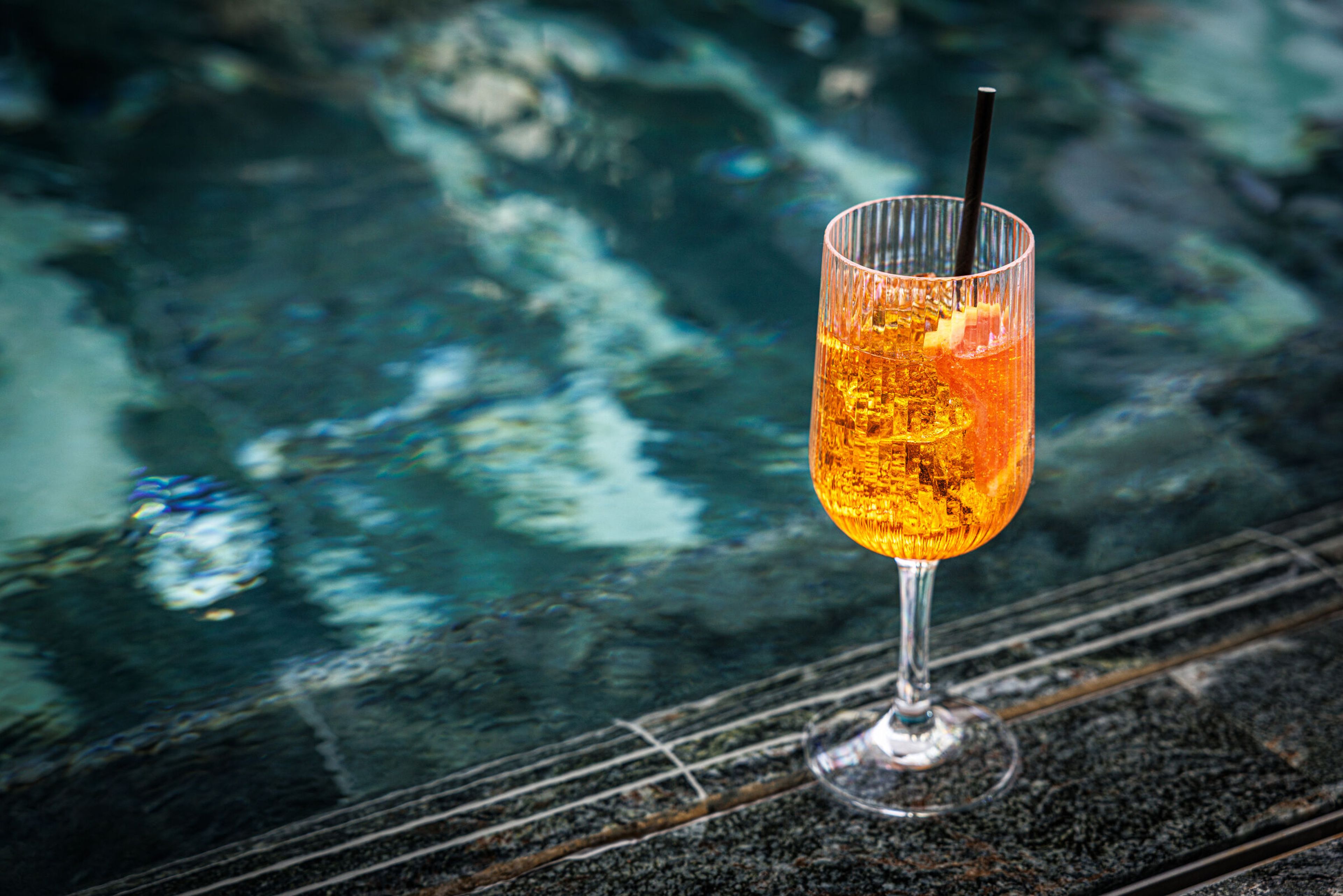 Räfflat vinglas med orange cocktail och citrusskiva placerat vid en pool.