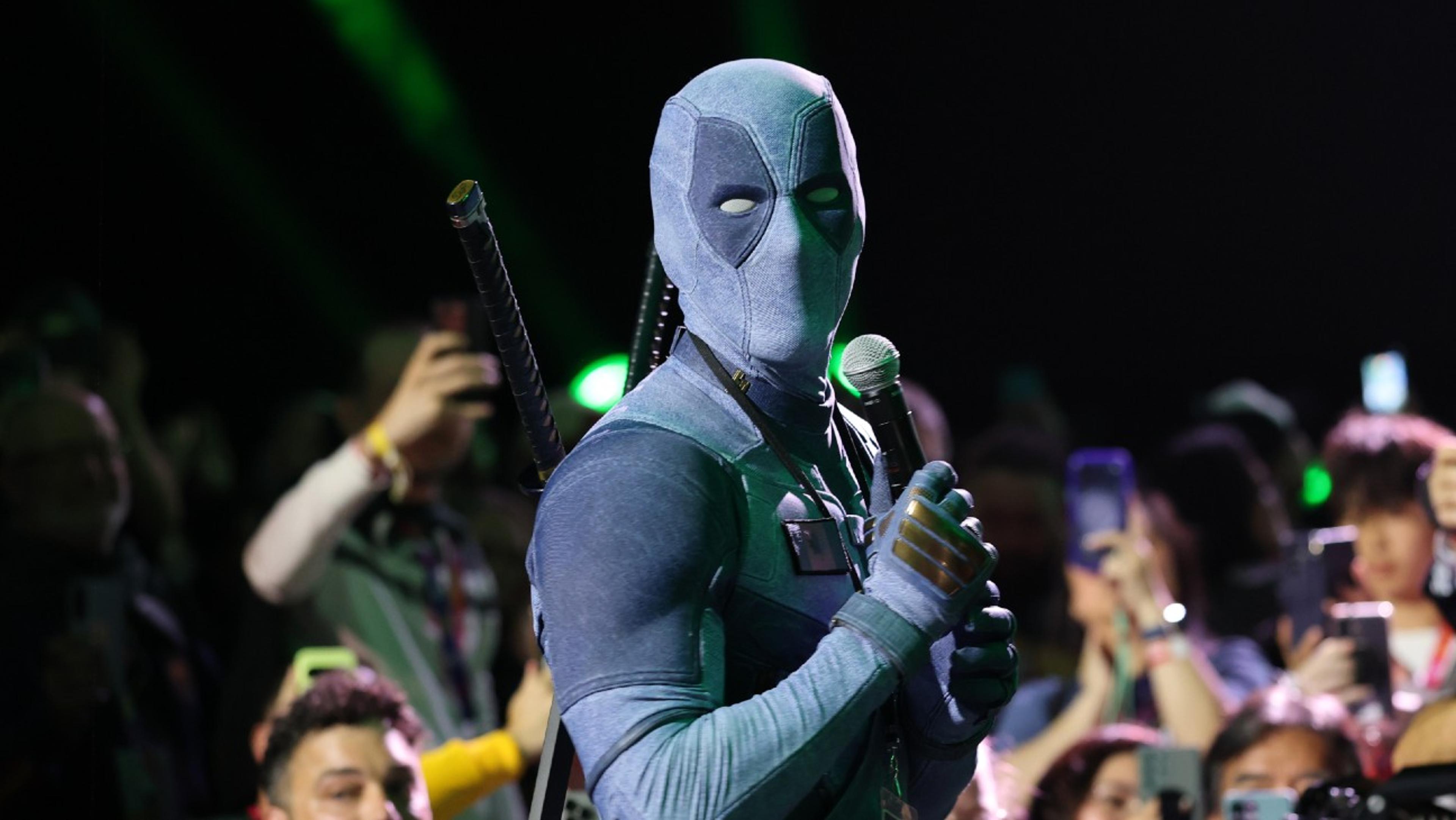 Ryan Reynolds in a grey-and-black Deadpool suit holding a microphone in the Hall H crowd, phones raised around him, at San Diego Comic-Con 2026.