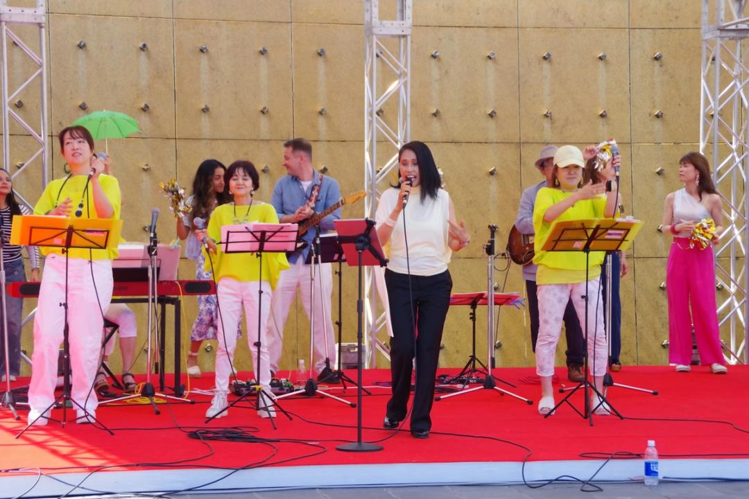 屋外ステージで、マイクを手に歌う女性たちと、楽器を演奏するバンドメンバー