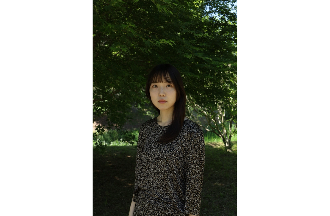 緑豊かな木々を背景に、木陰に立つ女性のポートレート。自然光が顔や周囲の葉にやわらかく差し込んでいる。
