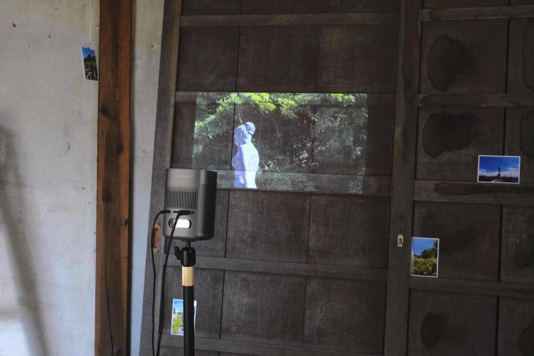 保泉エリ作品画像。人体彫刻が映る映像が、建物内の建具に投影されている。自立式スピーカーがある。屋外の写真が4枚空間の中に配置されている。