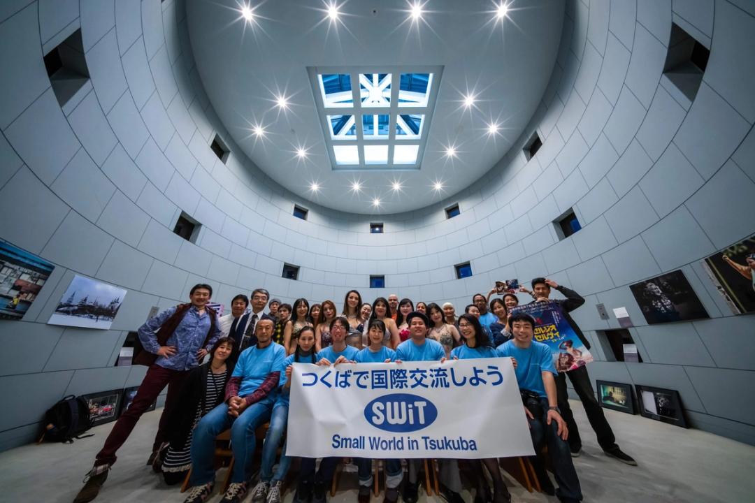 円形の展示空間で「つくばで国際交流しよう」と書かれた横断幕を掲げる、多国籍の参加者たちの集合写真