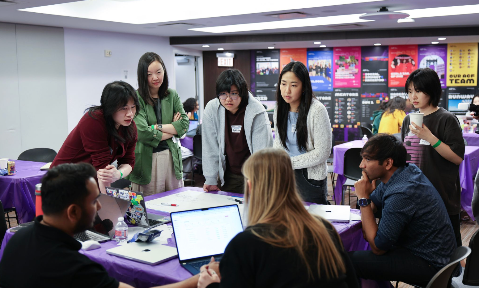 A group of students gathers around a table during a collaborative design workshop, leaning in to discuss ideas while laptops, notes, and a whiteboard are spread across the table. Additional participants work in the background, with colorful event posters lining the wall, creating an energetic classroom and studio atmosphere.