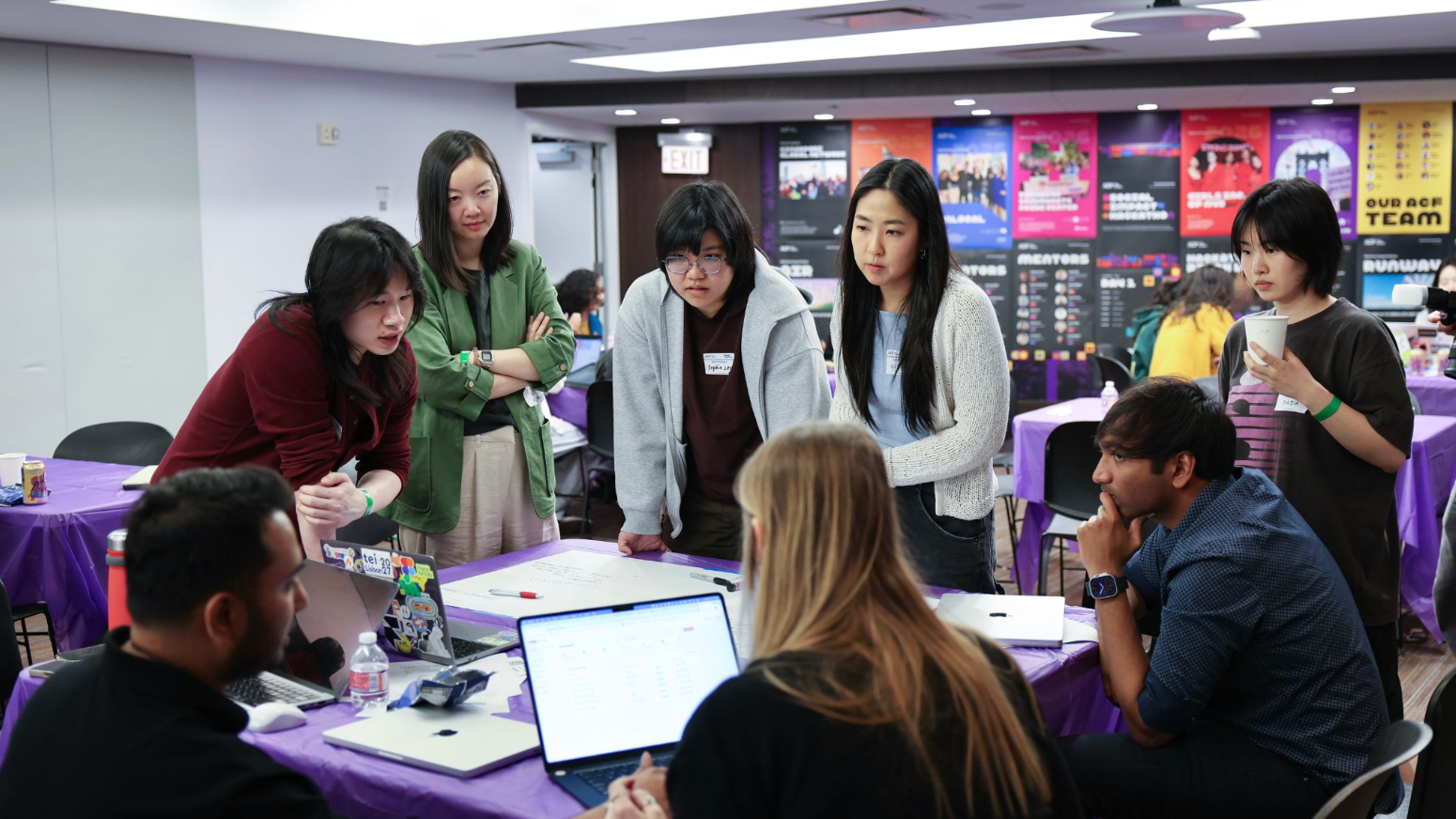 A group of students gathers around a table during a collaborative design workshop, leaning in to discuss ideas while laptops, notes, and a whiteboard are spread across the table. Additional participants work in the background, with colorful event posters lining the wall, creating an energetic classroom and studio atmosphere.