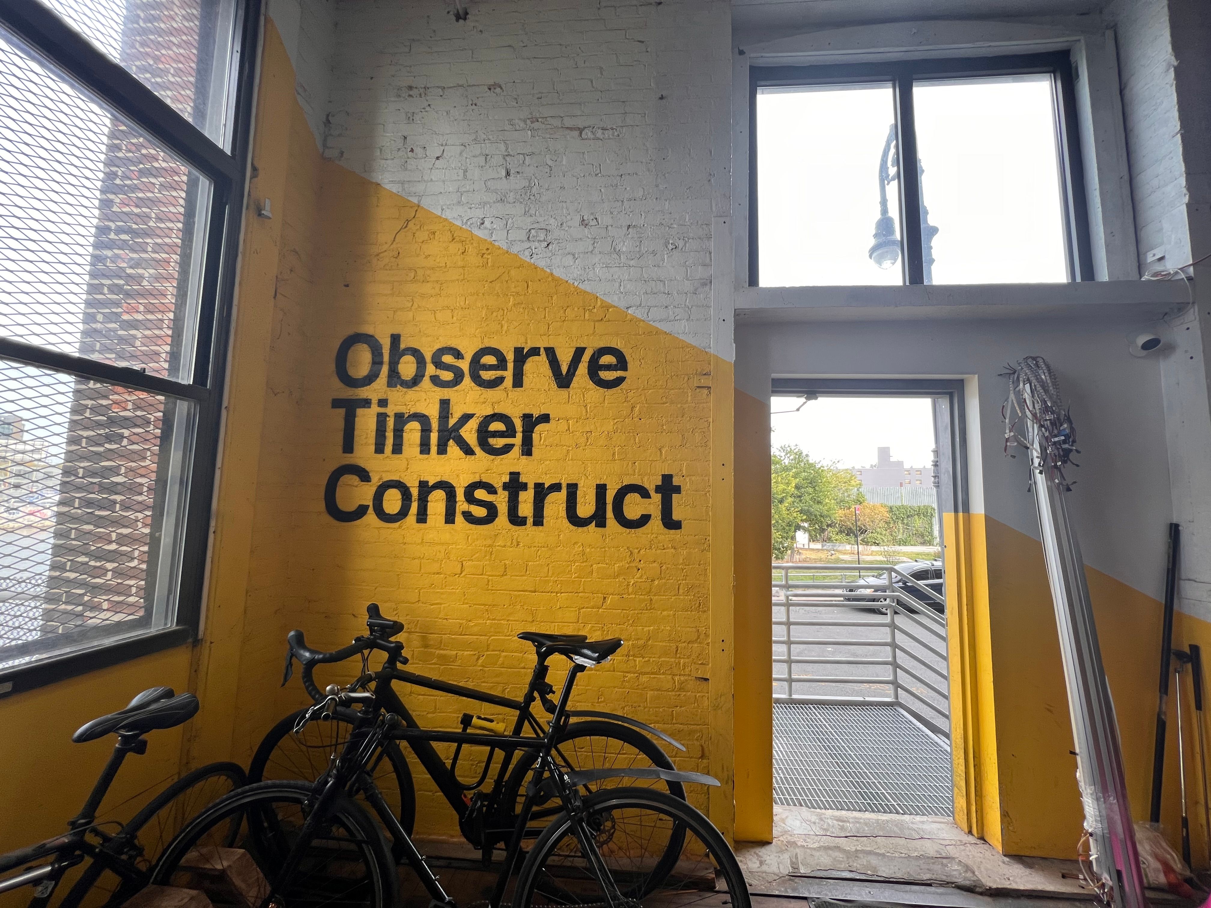 Bikes in a bike stand in front of a bright yellow wall with the words Observe Tinker Construct written on it in bold black typeface.