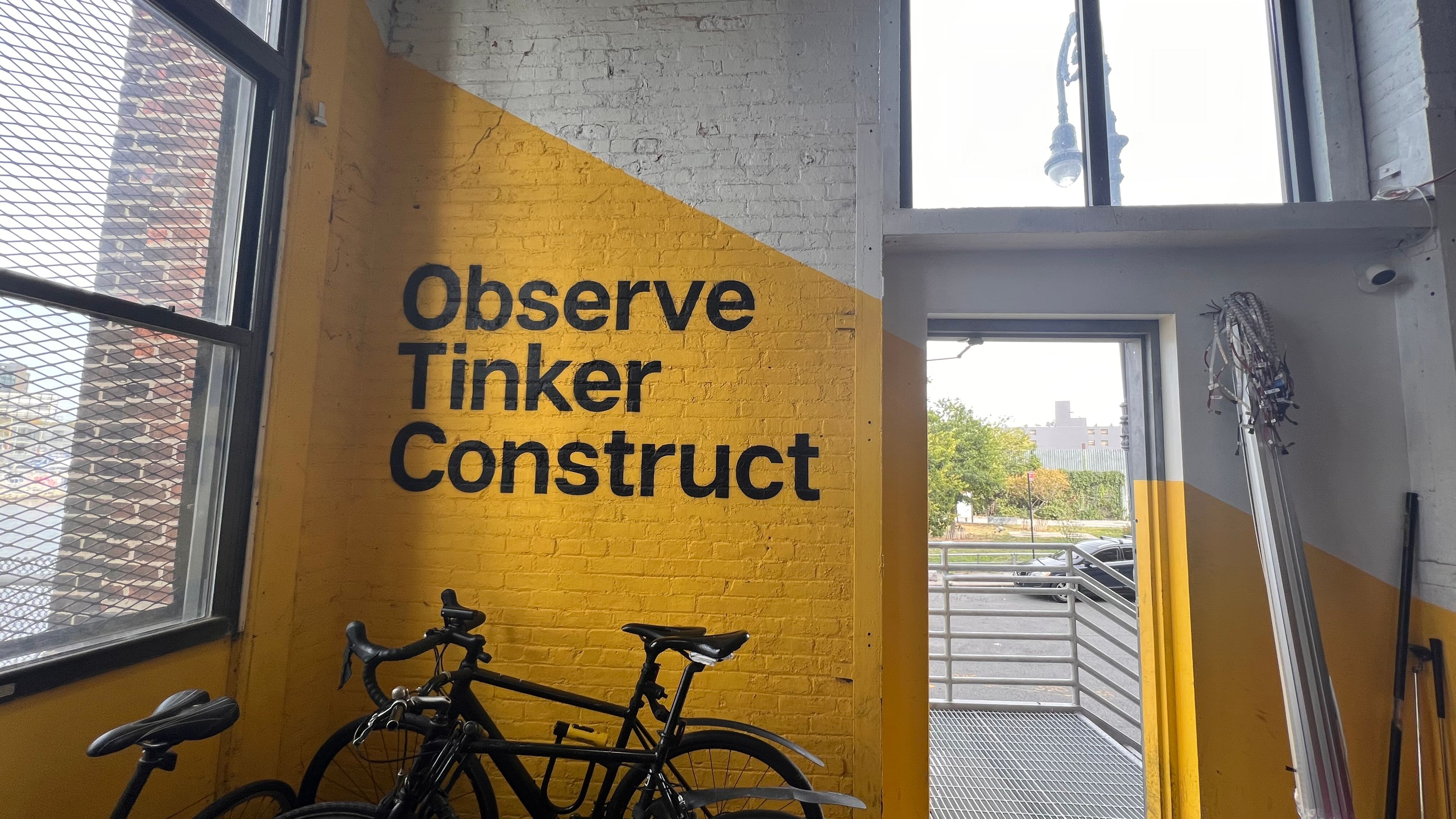 Bikes in a bike stand in front of a bright yellow wall with the words Observe Tinker Construct written on it in bold black typeface.