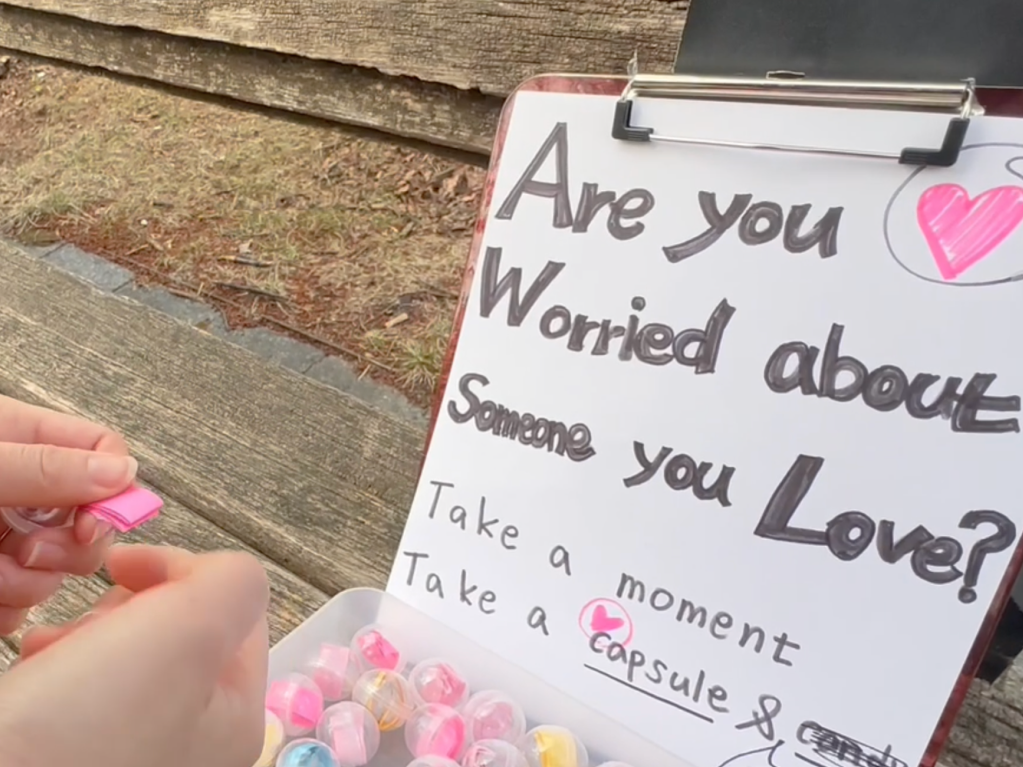 A person selects a small pink capsule from a container beside a handwritten sign encouraging people to take a moment if they are worried about someone they love.