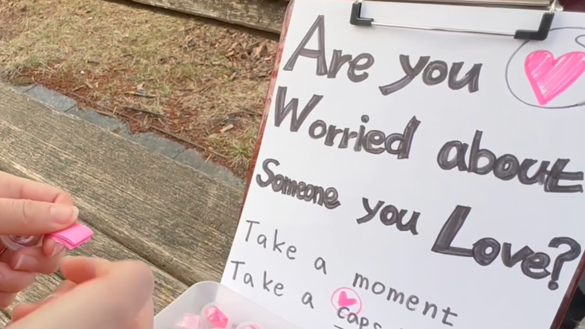A person selects a small pink capsule from a container beside a handwritten sign encouraging people to take a moment if they are worried about someone they love.