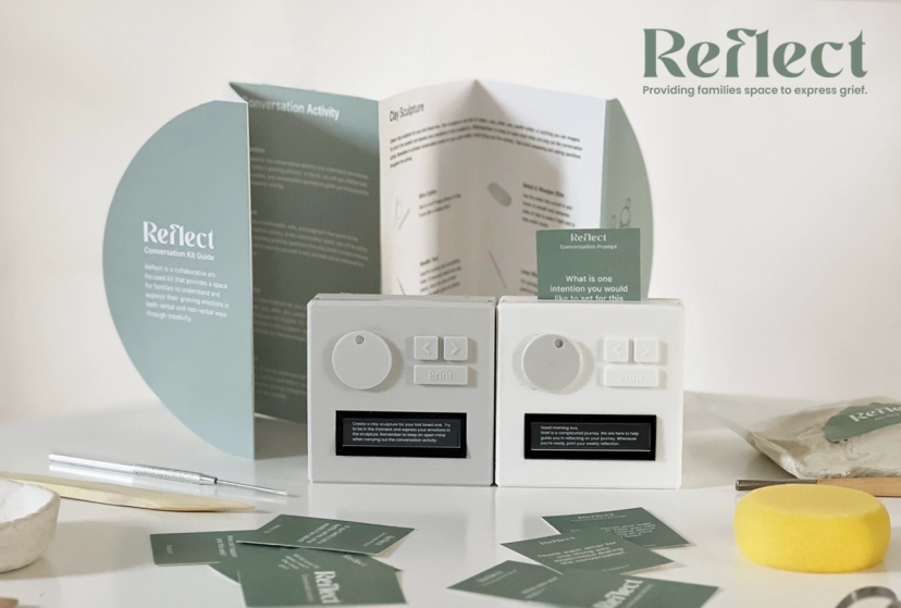 Presentation of Reflect, a conversation kit designed to help families express grief through creative activities. The display includes a folded instruction guide, two small white interactive devices with knobs, buttons, and digital screens, and scattered conversation prompt cards in muted green. A prompt card is inserted into one of the devices, while clay sculpting instructions are visible in the open guide behind them. Additional materials, including sculpting tools, clay, and a yellow sponge, are arranged on the table. The Reflect logo and tagline, “Providing families space to express grief,” appear in the upper right against a soft, neutral background.