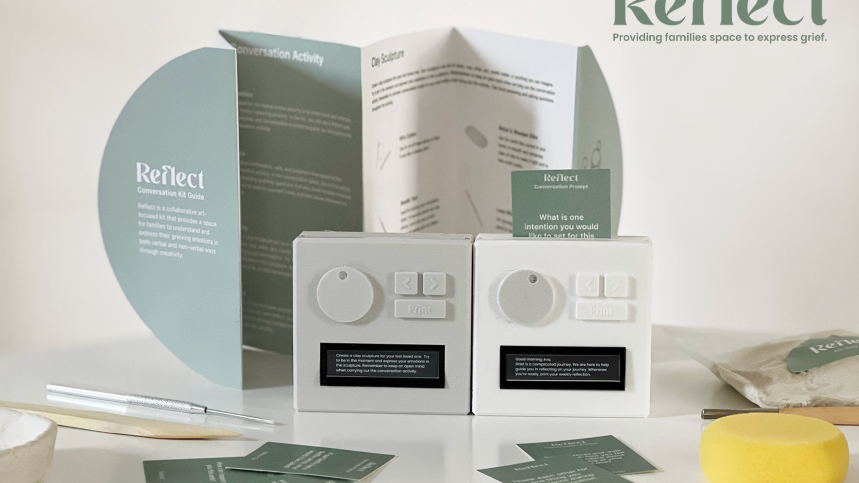 Presentation of Reflect, a conversation kit designed to help families express grief through creative activities. The display includes a folded instruction guide, two small white interactive devices with knobs, buttons, and digital screens, and scattered conversation prompt cards in muted green. A prompt card is inserted into one of the devices, while clay sculpting instructions are visible in the open guide behind them. Additional materials, including sculpting tools, clay, and a yellow sponge, are arranged on the table. The Reflect logo and tagline, “Providing families space to express grief,” appear in the upper right against a soft, neutral background.