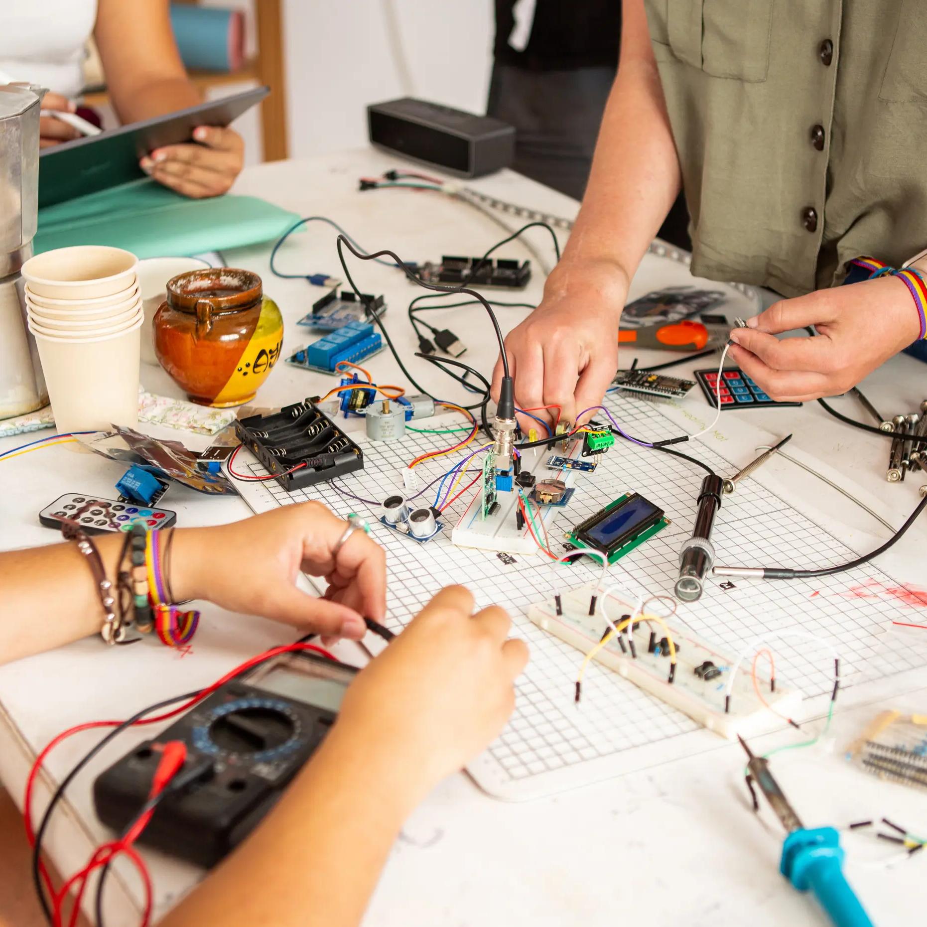 A group of people working on an electronics project at a cluttered table filled with circuit boards, wires, resistors, and other electronic components.
