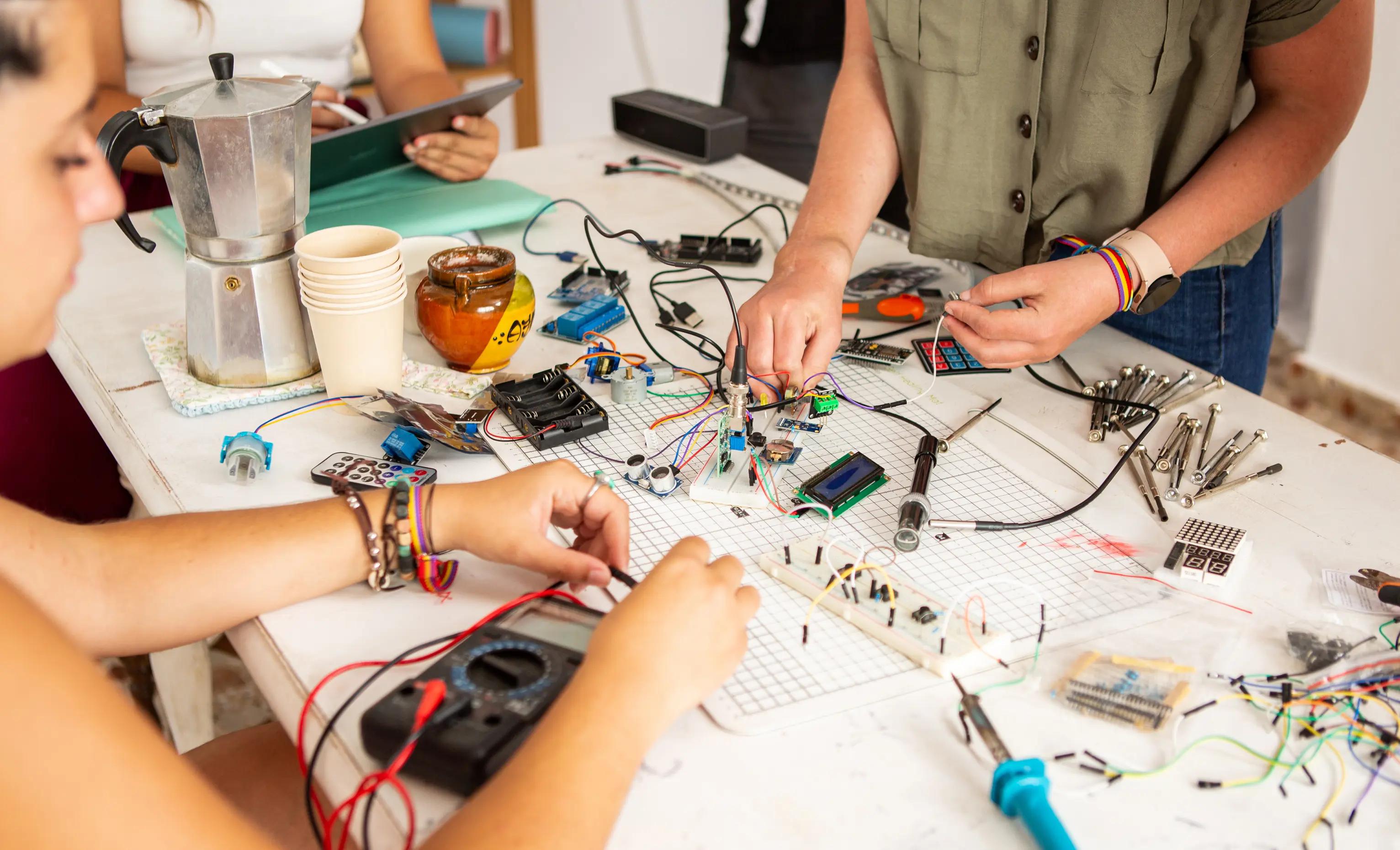 A group of people working on an electronics project at a cluttered table filled with circuit boards, wires, resistors, and other electronic components.