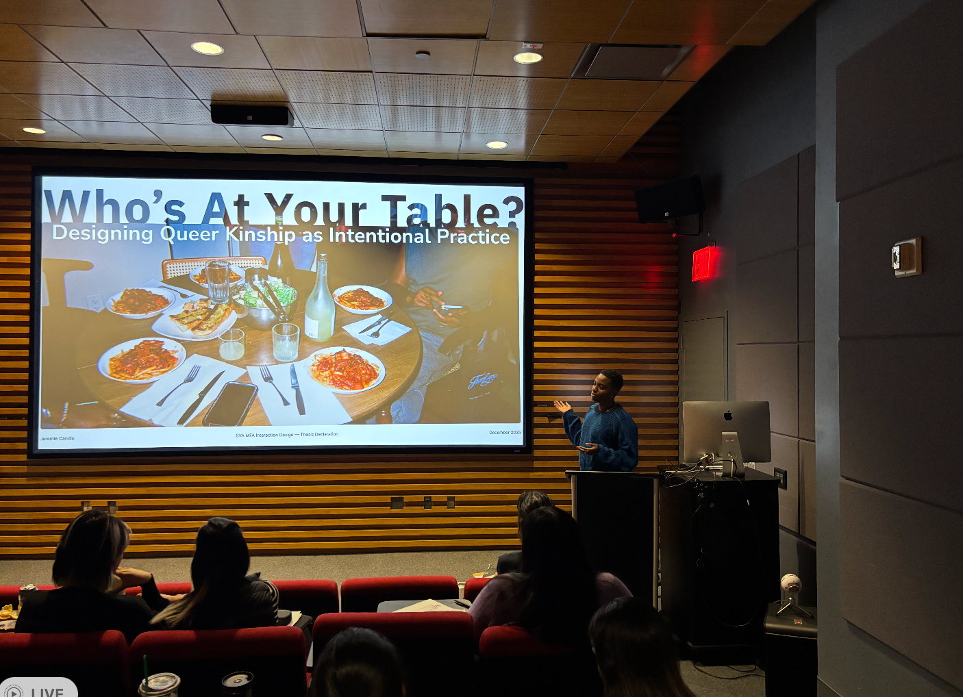 Student presenting their work in the SVA theater. The words Who's At Your Table? and a photo of a dinner table are projected on a large screen as he speaks. 