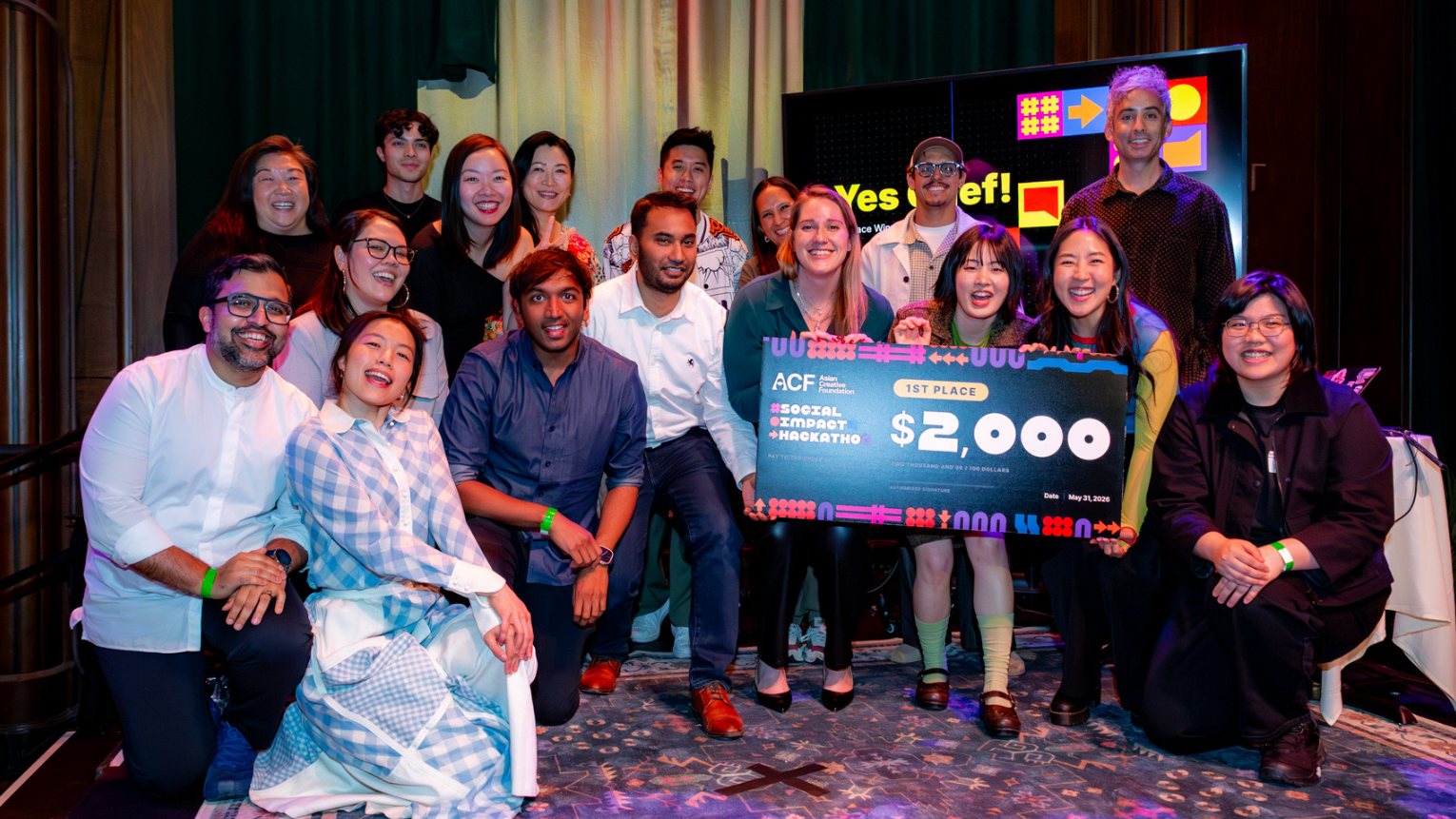 A large group of smiling participants poses together on stage after a design competition, with the winning team holding an oversized check for $2,000 labeled "1st Place" from the ACF Social Impact Hackathon. A presentation screen in the background displays colorful graphics and the words "Yes Chef!", capturing a celebratory moment following the award presentation.