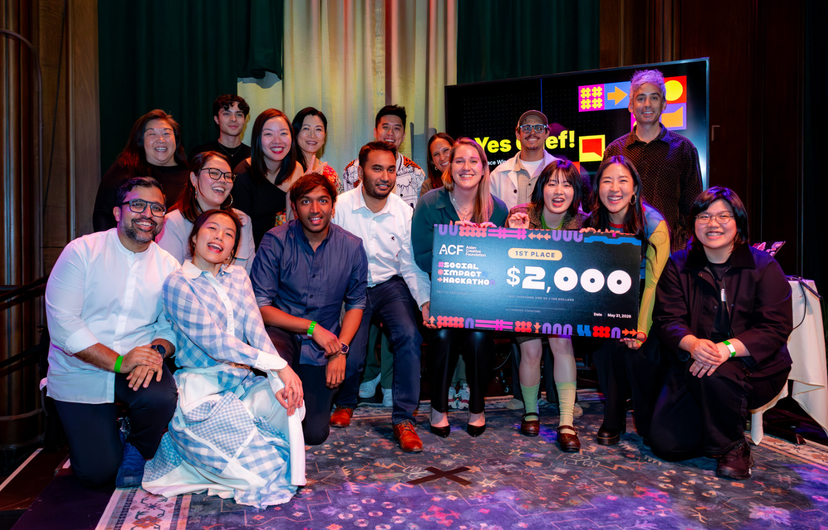 A large group of smiling participants poses together on stage after a design competition, with the winning team holding an oversized check for $2,000 labeled "1st Place" from the ACF Social Impact Hackathon. A presentation screen in the background displays colorful graphics and the words "Yes Chef!", capturing a celebratory moment following the award presentation.