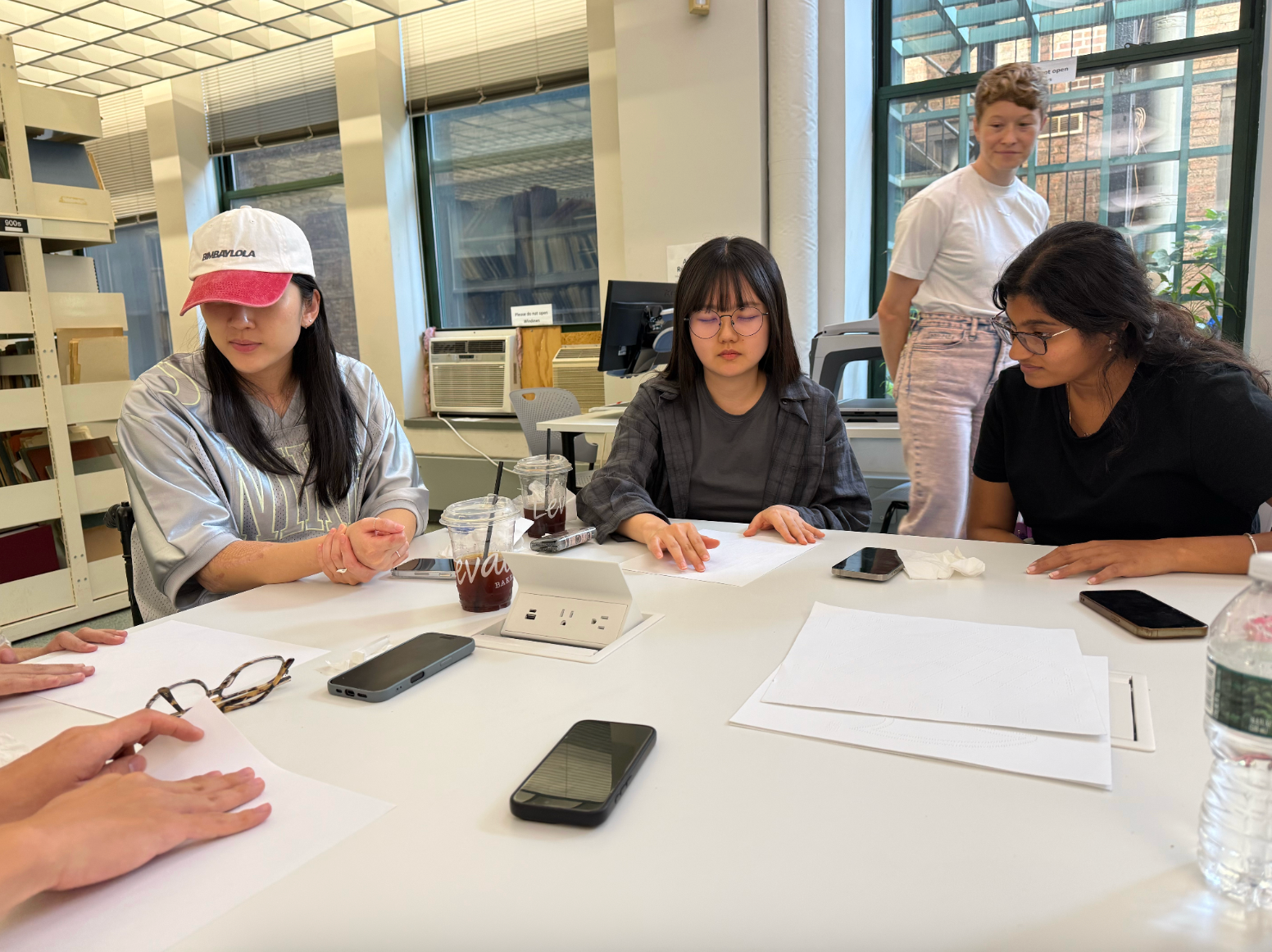 A group of students gathers around a large table during a collaborative workshop in a bright classroom. Three students are seated, focused on blank sheets of paper in front of them, while another student stands nearby observing the discussion. The table is scattered with notebooks, smartphones, iced coffee cups, tissues, and a water bottle. Large windows, bookshelves, and computer workstations in the background create an open studio environment.