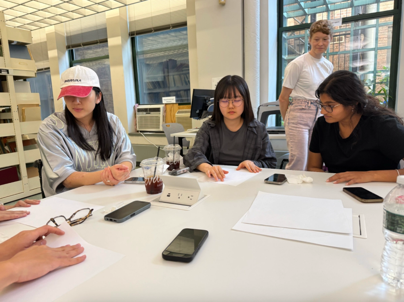 A group of students gathers around a large table during a collaborative workshop in a bright classroom. Three students are seated, focused on blank sheets of paper in front of them, while another student stands nearby observing the discussion. The table is scattered with notebooks, smartphones, iced coffee cups, tissues, and a water bottle. Large windows, bookshelves, and computer workstations in the background create an open studio environment.