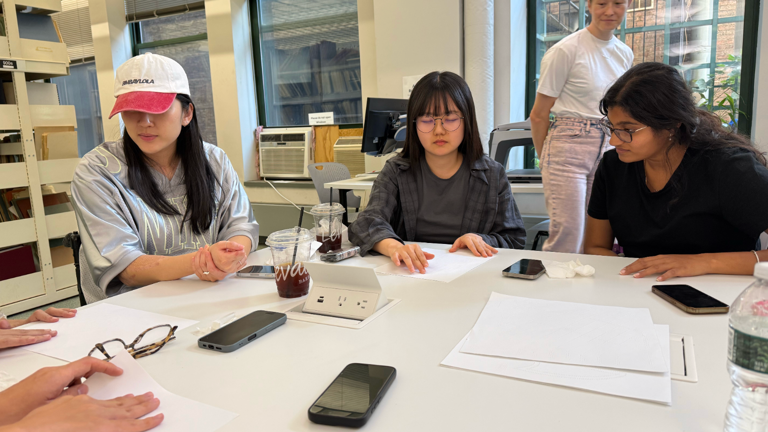 A group of students gathers around a large table during a collaborative workshop in a bright classroom. Three students are seated, focused on blank sheets of paper in front of them, while another student stands nearby observing the discussion. The table is scattered with notebooks, smartphones, iced coffee cups, tissues, and a water bottle. Large windows, bookshelves, and computer workstations in the background create an open studio environment.