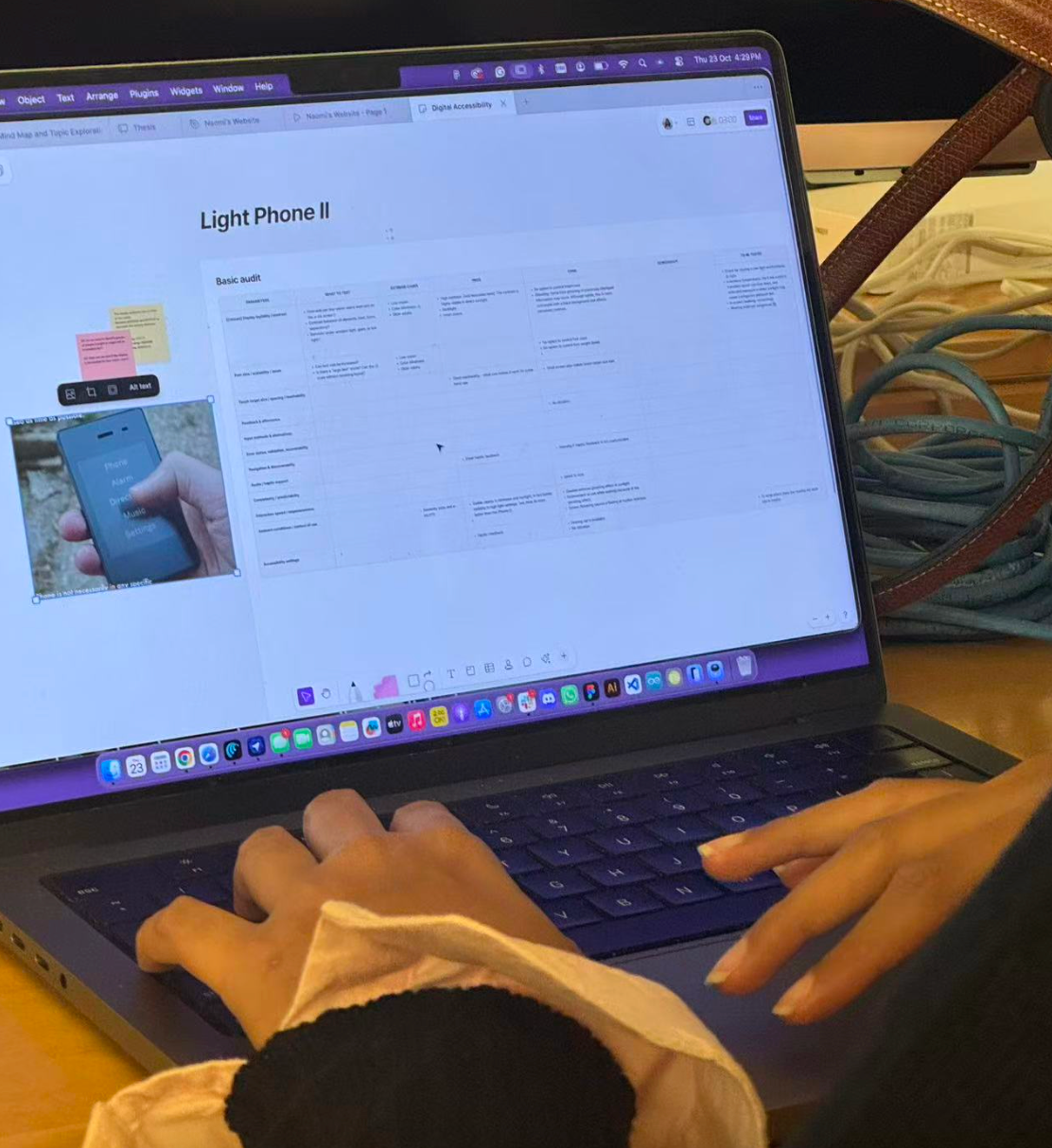 Close-up of a person working on a laptop displaying a digital accessibility audit for the Light Phone II in a collaborative design workspace. The screen shows a whiteboard with a table of audit findings, sticky notes, and an image of the phone interface. The person's hands rest on the keyboard and trackpad, with coiled cables and a desktop monitor visible in the background.