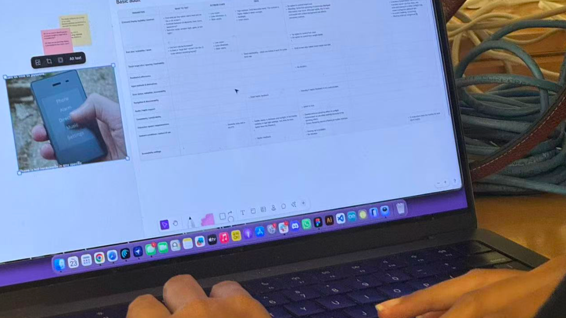 Close-up of a person working on a laptop displaying a digital accessibility audit for the Light Phone II in a collaborative design workspace. The screen shows a whiteboard with a table of audit findings, sticky notes, and an image of the phone interface. The person's hands rest on the keyboard and trackpad, with coiled cables and a desktop monitor visible in the background.