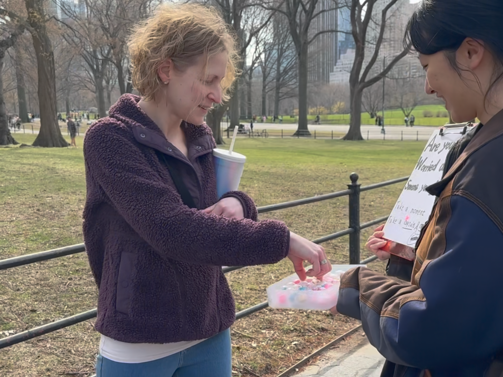 Two people stand together in a public park on a sunny day beside a wellness outreach display. One person holds a container filled with colorful capsules while the other reaches in to select one. A partially visible sign invites participants to reflect on concerns about someone they care about and to take a capsule. Trees, pathways, and city buildings in the background create an open, community-centered setting. The scene conveys connection, curiosity, and engagement with an interactive mental health and support initiative.