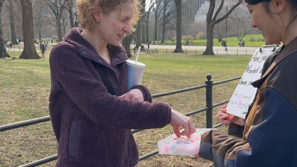 Two people stand together in a public park on a sunny day beside a wellness outreach display. One person holds a container filled with colorful capsules while the other reaches in to select one. A partially visible sign invites participants to reflect on concerns about someone they care about and to take a capsule. Trees, pathways, and city buildings in the background create an open, community-centered setting. The scene conveys connection, curiosity, and engagement with an interactive mental health and support initiative.