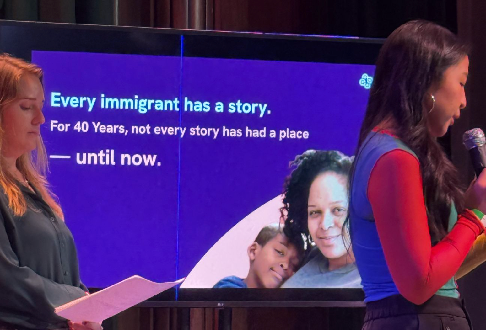 Two presenters stand on stage in front of a large presentation screen. One person reads from a microphone while the other holds notes. The slide behind them reads, "Every immigrant has a story. For 40 Years, not every story has had a place—until now," alongside a photo of a parent and child, highlighting a storytelling and community-centered presentation.