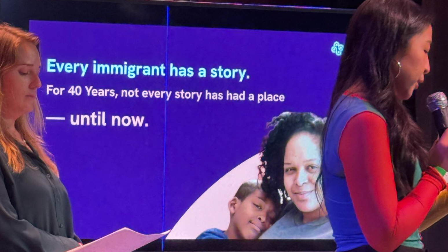 Two presenters stand on stage in front of a large presentation screen. One person reads from a microphone while the other holds notes. The slide behind them reads, "Every immigrant has a story. For 40 Years, not every story has had a place—until now," alongside a photo of a parent and child, highlighting a storytelling and community-centered presentation.