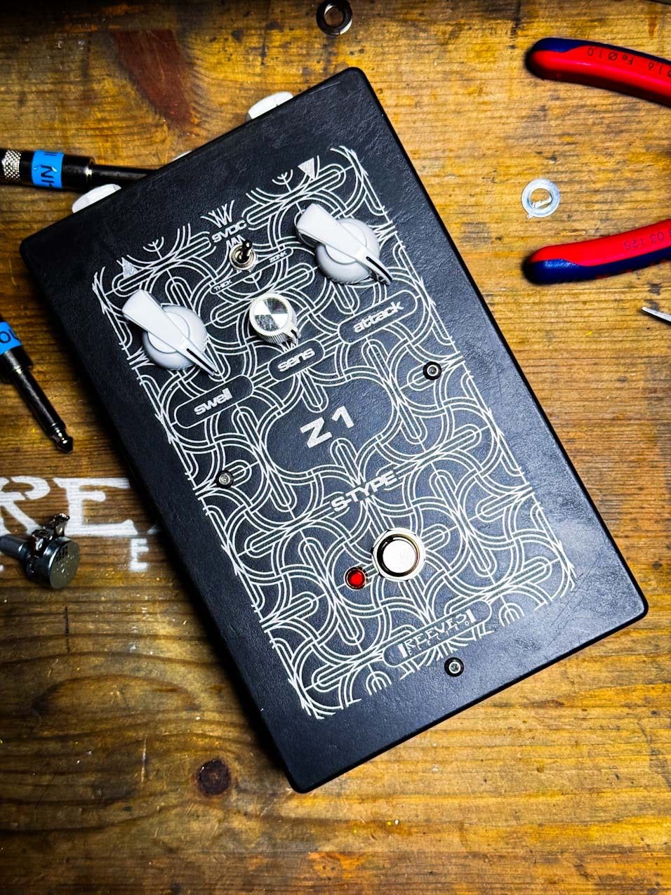 Reeves Electro Z1 fuzz on a wooden workbench among pliers and tools, black acid-etched enclosure with three white knobs and a red LED