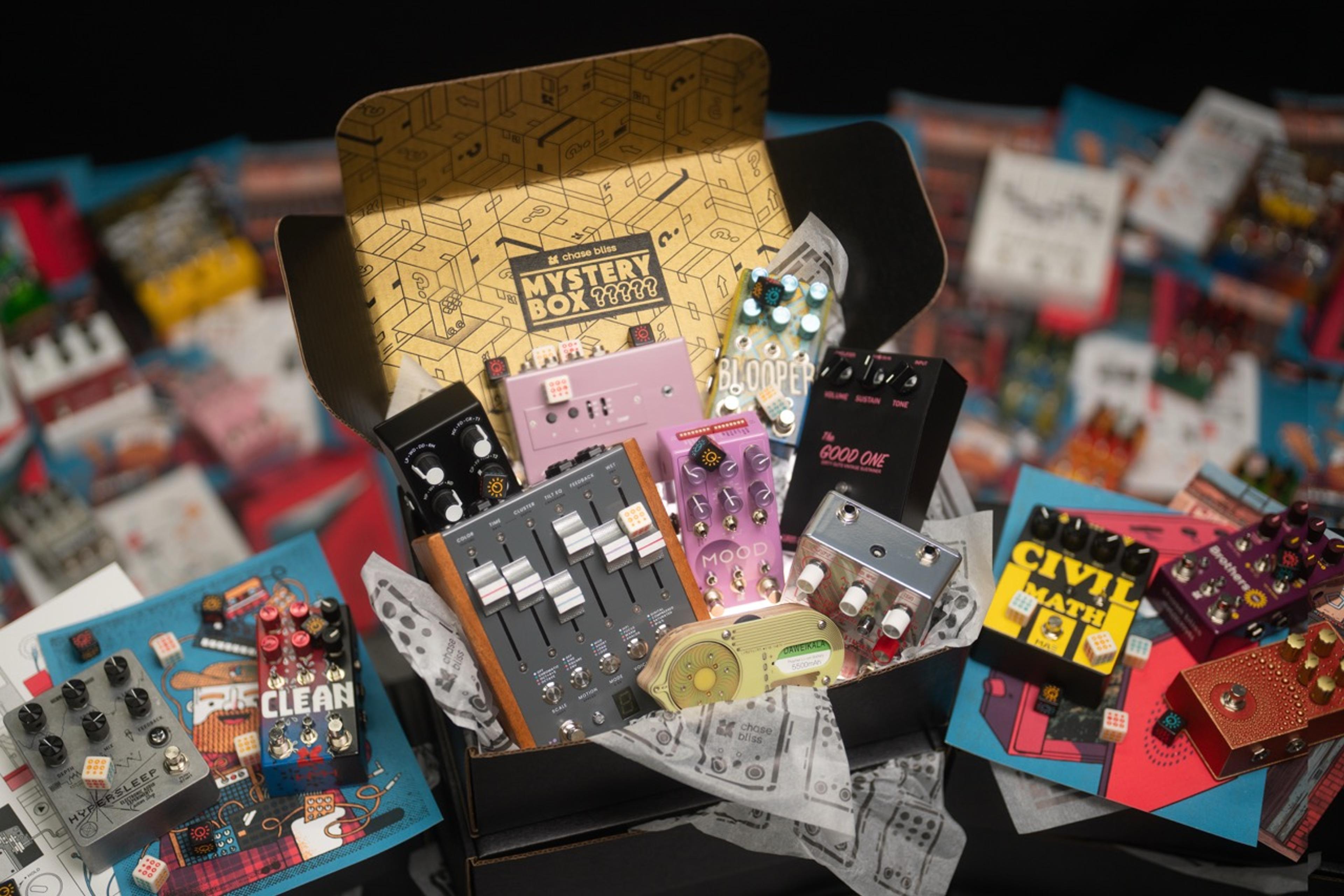 Open Chase Bliss Mystery Box with a gold printed lid, filled with Chase Bliss pedals, dice and artist prints spread around it
