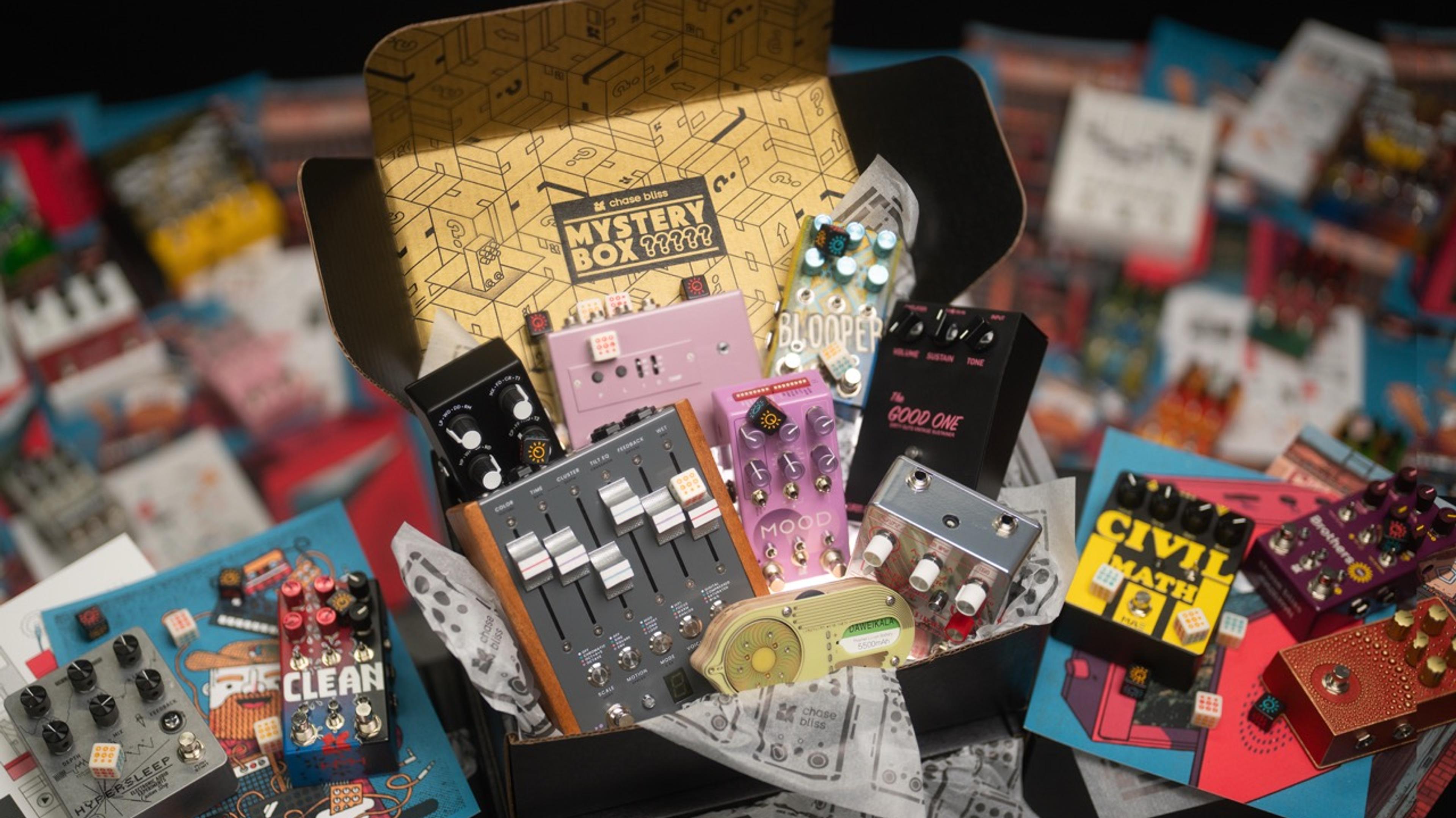 Open Chase Bliss Mystery Box with a gold printed lid, filled with Chase Bliss pedals, dice and artist prints spread around it