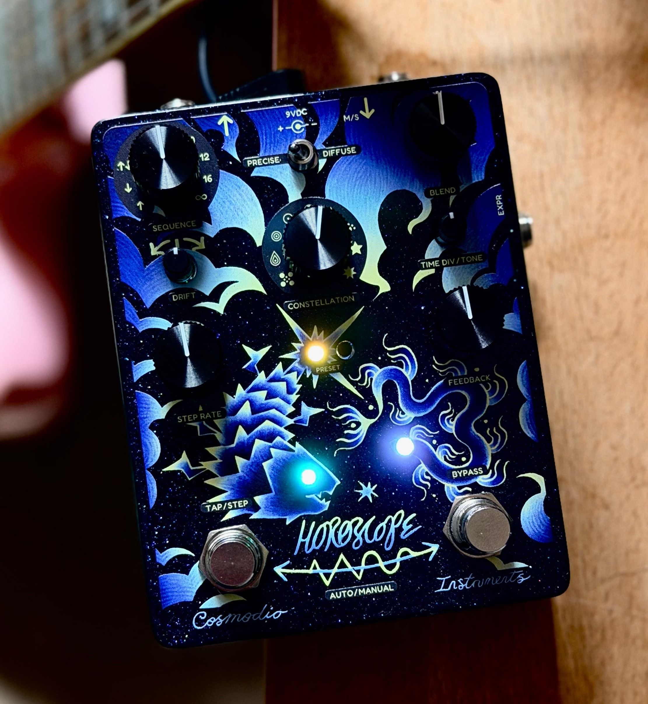 Cosmodio Horoscope leaning against a guitar neck on warm wood, preset and status LEDs lit through the blue fish-and-serpent artwork