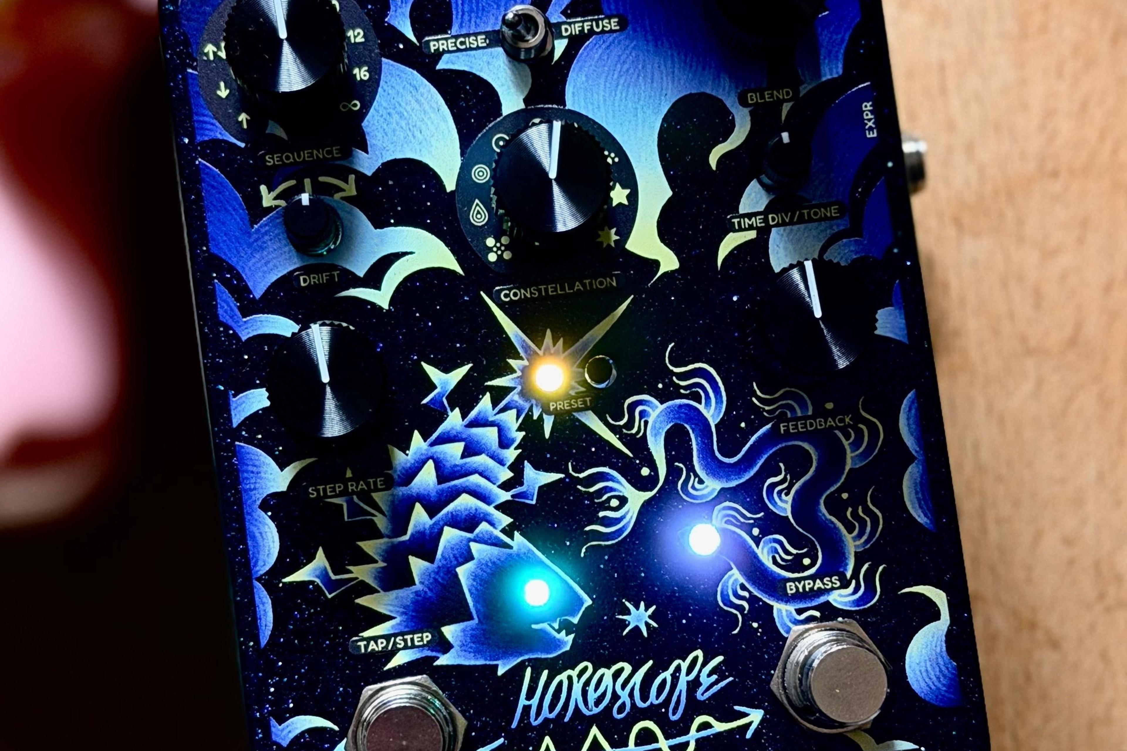 Cosmodio Horoscope leaning against a guitar neck on warm wood, preset and status LEDs lit through the blue fish-and-serpent artwork