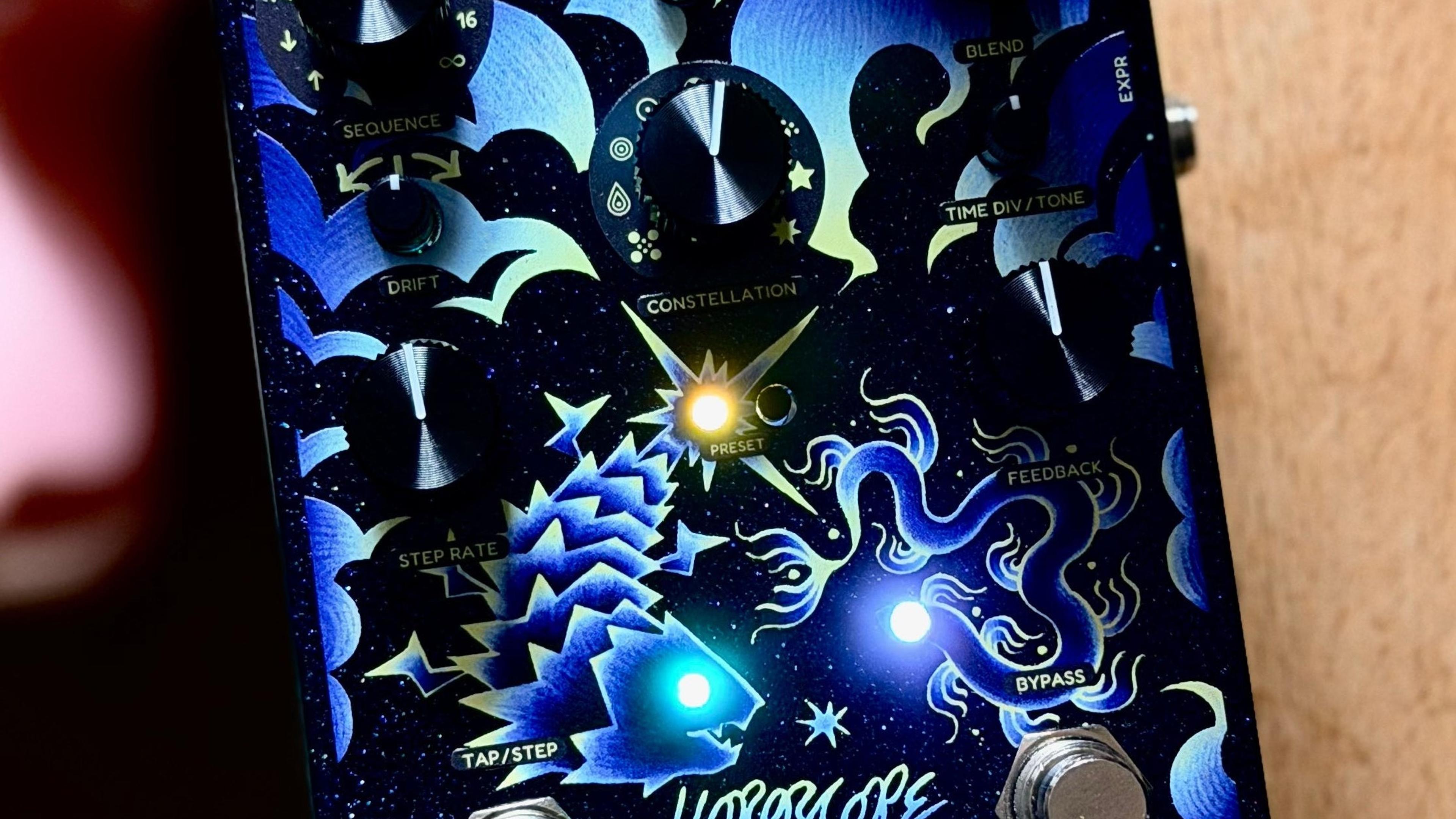 Cosmodio Horoscope leaning against a guitar neck on warm wood, preset and status LEDs lit through the blue fish-and-serpent artwork