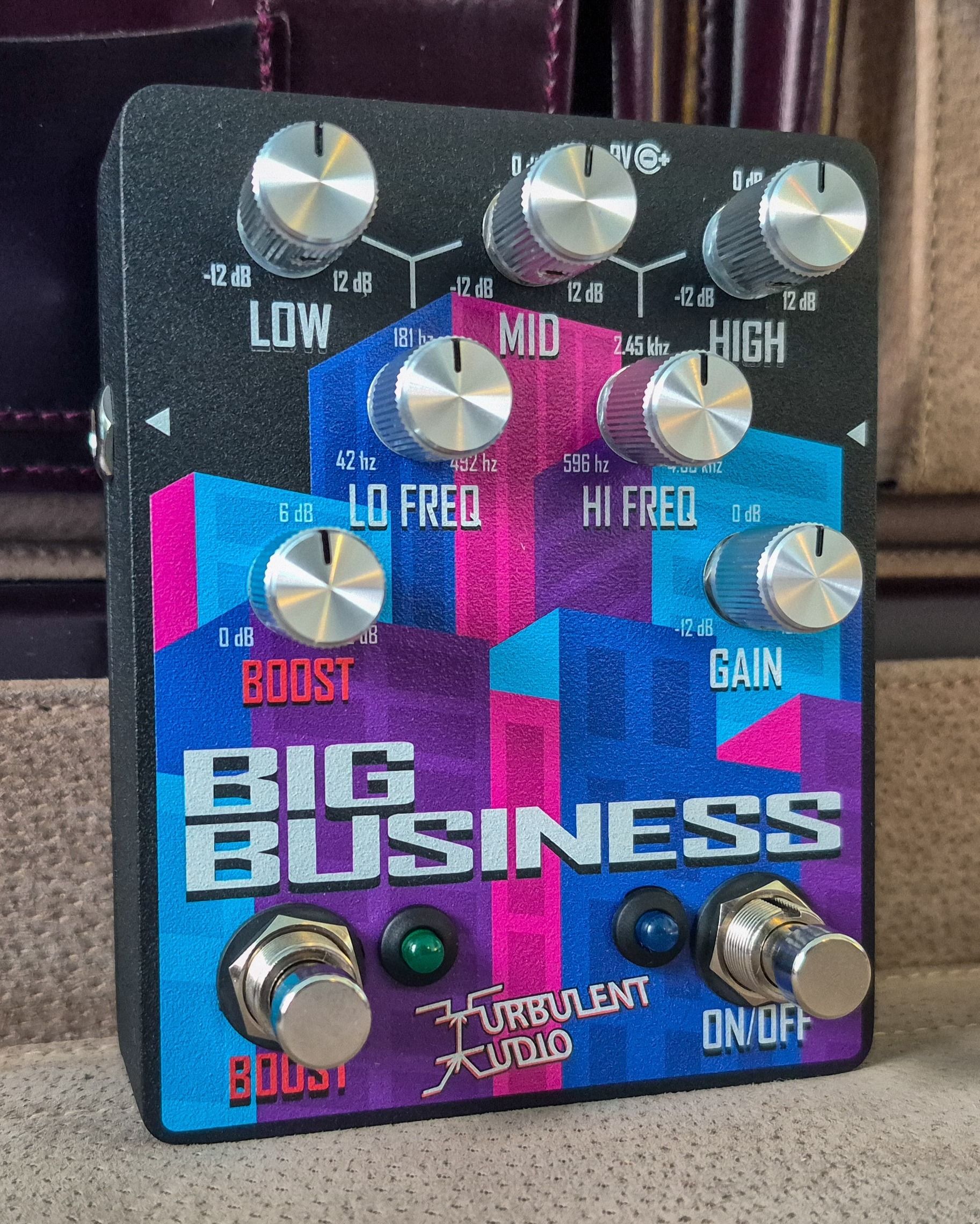 Turbulent Audio Big Business pedal standing on a beige leather surface in front of a brown leather briefcase, skyline graphic and eight silver knobs