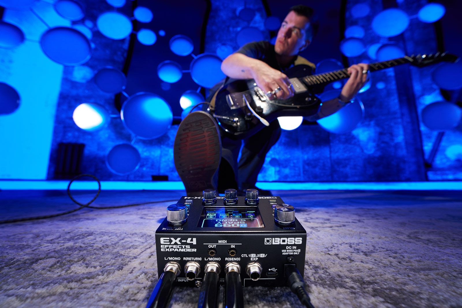 Boss EX-4 Effects Expander on a studio floor with a guitarist playing in a blue-lit room behind it