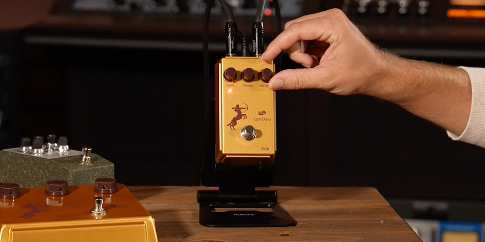 Warm Audio Centavo Mod gold overdrive pedal on a studio stand with cables and a hand adjusting Output