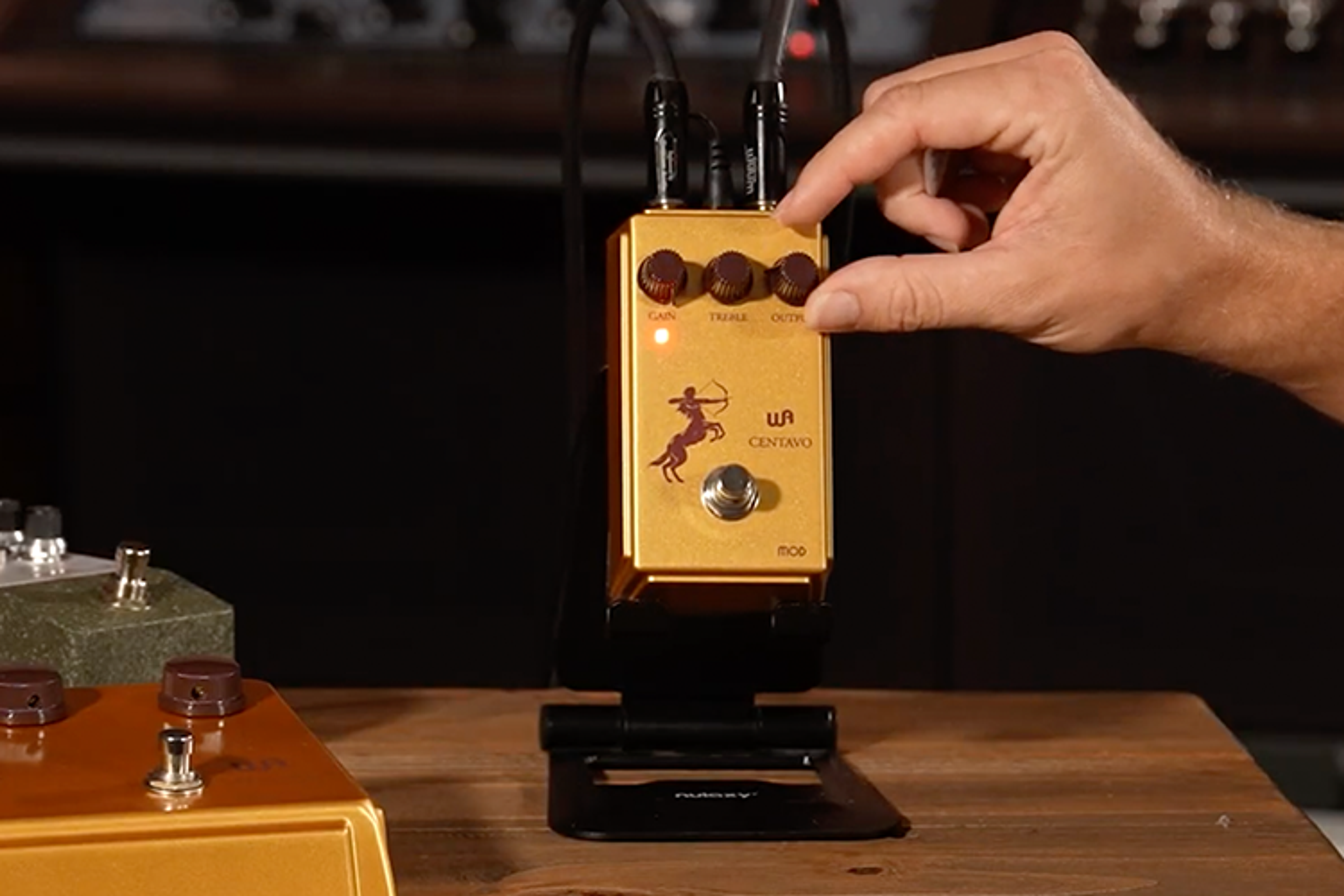 Warm Audio Centavo Mod gold overdrive pedal on a studio stand with cables and a hand adjusting Output