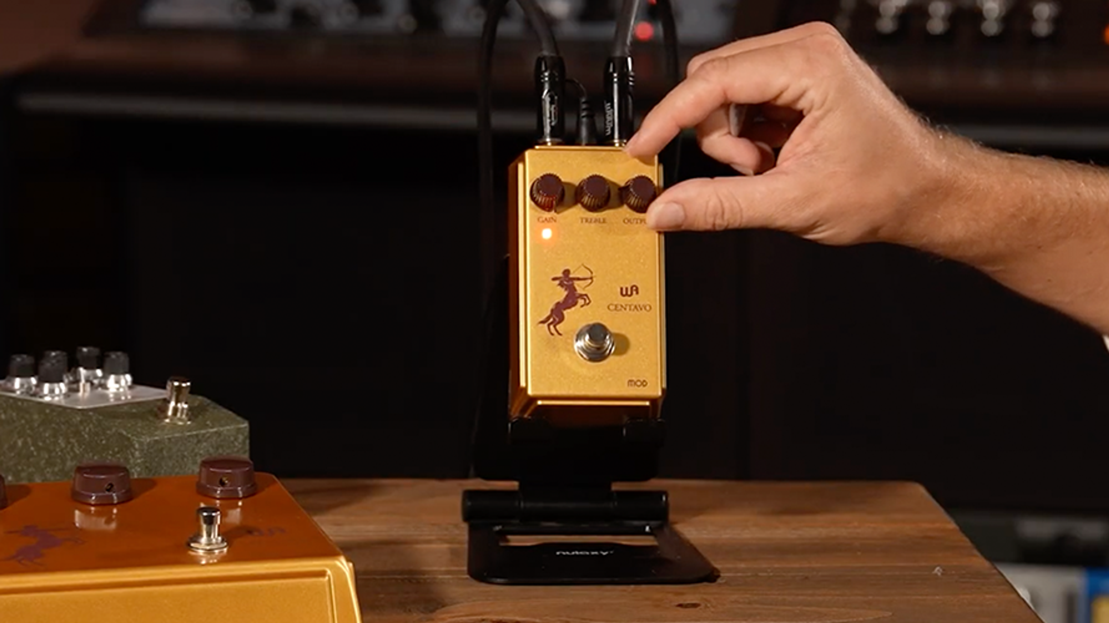 Warm Audio Centavo Mod gold overdrive pedal on a studio stand with cables and a hand adjusting Output