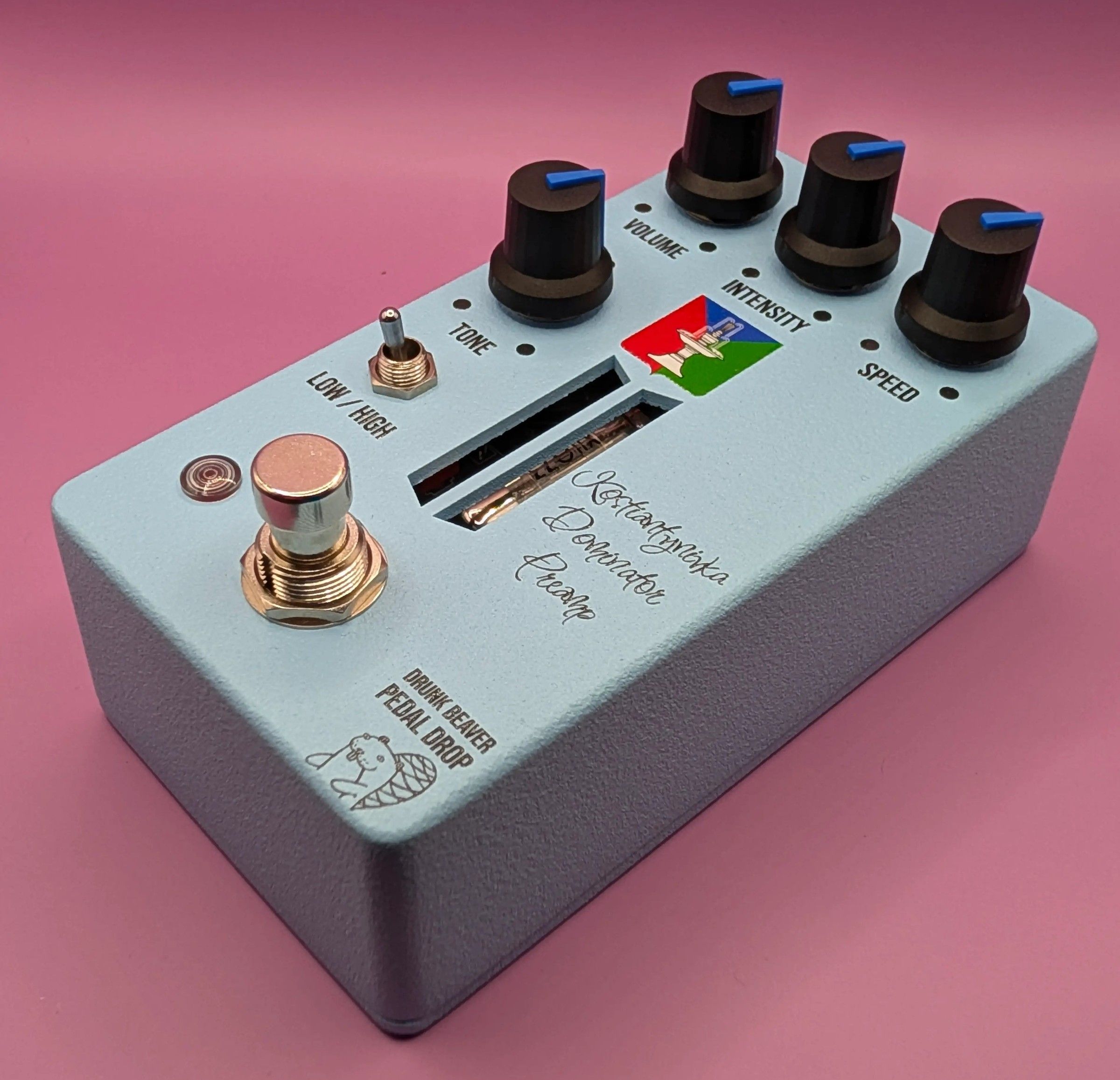 Drunk Beaver Kostiantynivka Dominator Preamp, three-quarter view on a pink backdrop, pale blue enclosure with four black knobs, Low/High toggle and copper footswitch