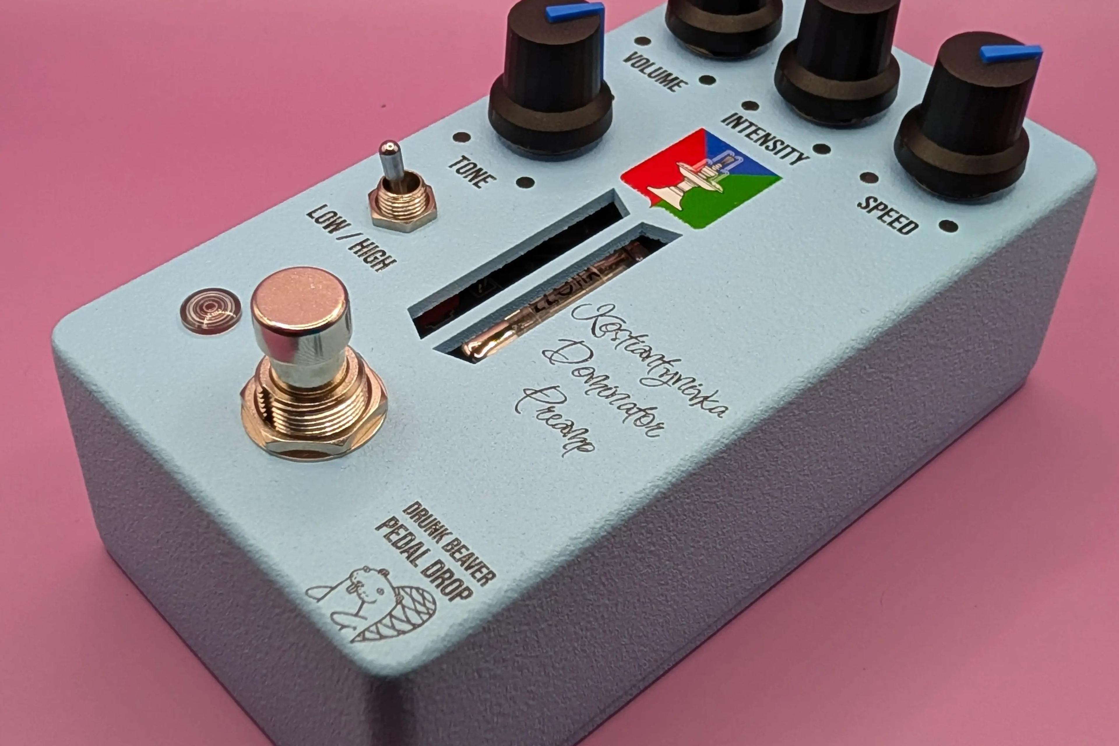 Drunk Beaver Kostiantynivka Dominator Preamp, three-quarter view on a pink backdrop, pale blue enclosure with four black knobs, Low/High toggle and copper footswitch