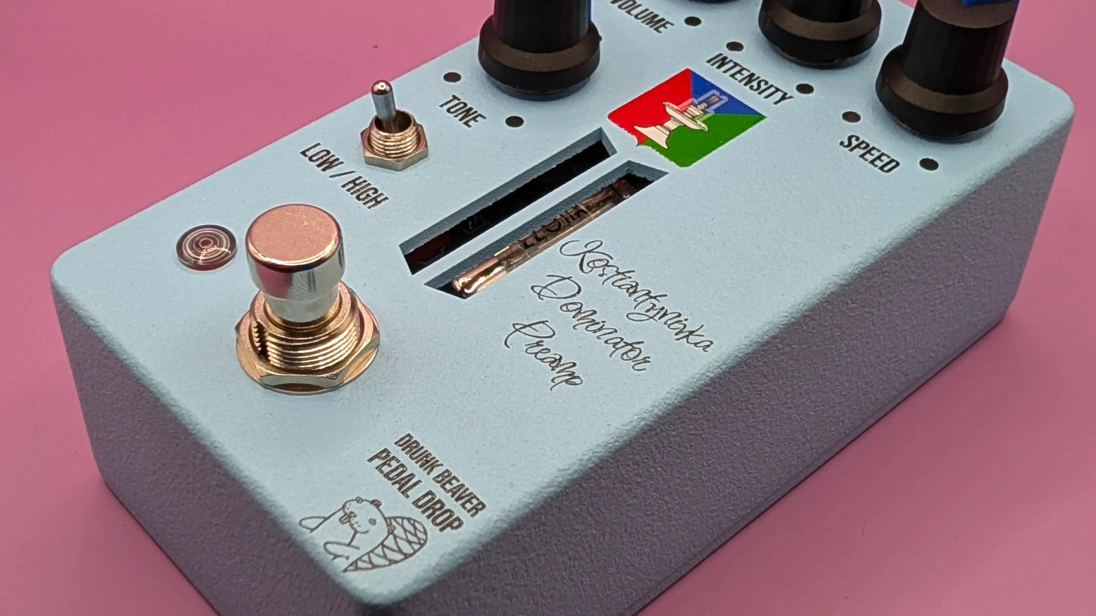 Drunk Beaver Kostiantynivka Dominator Preamp, three-quarter view on a pink backdrop, pale blue enclosure with four black knobs, Low/High toggle and copper footswitch