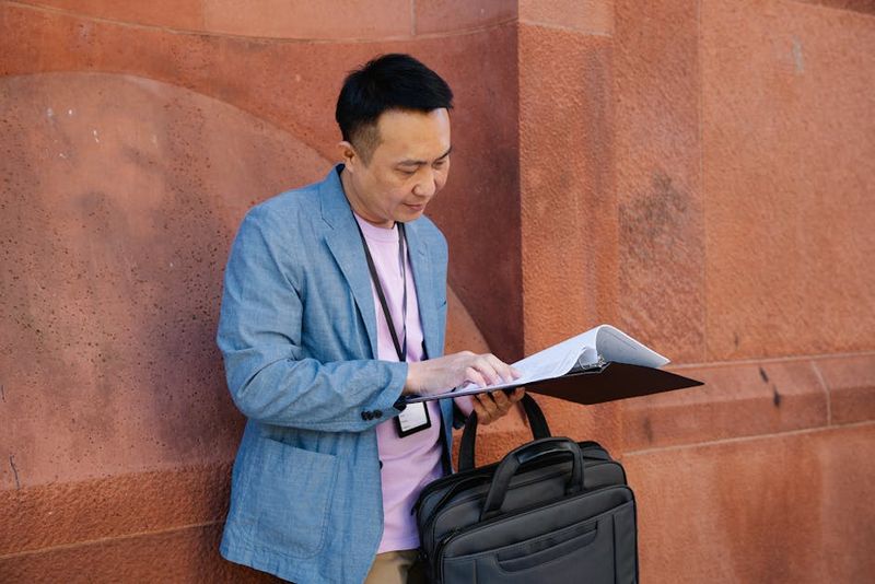 Asian man in a blue suit jacket reading documents from a binder while standing outdoors.